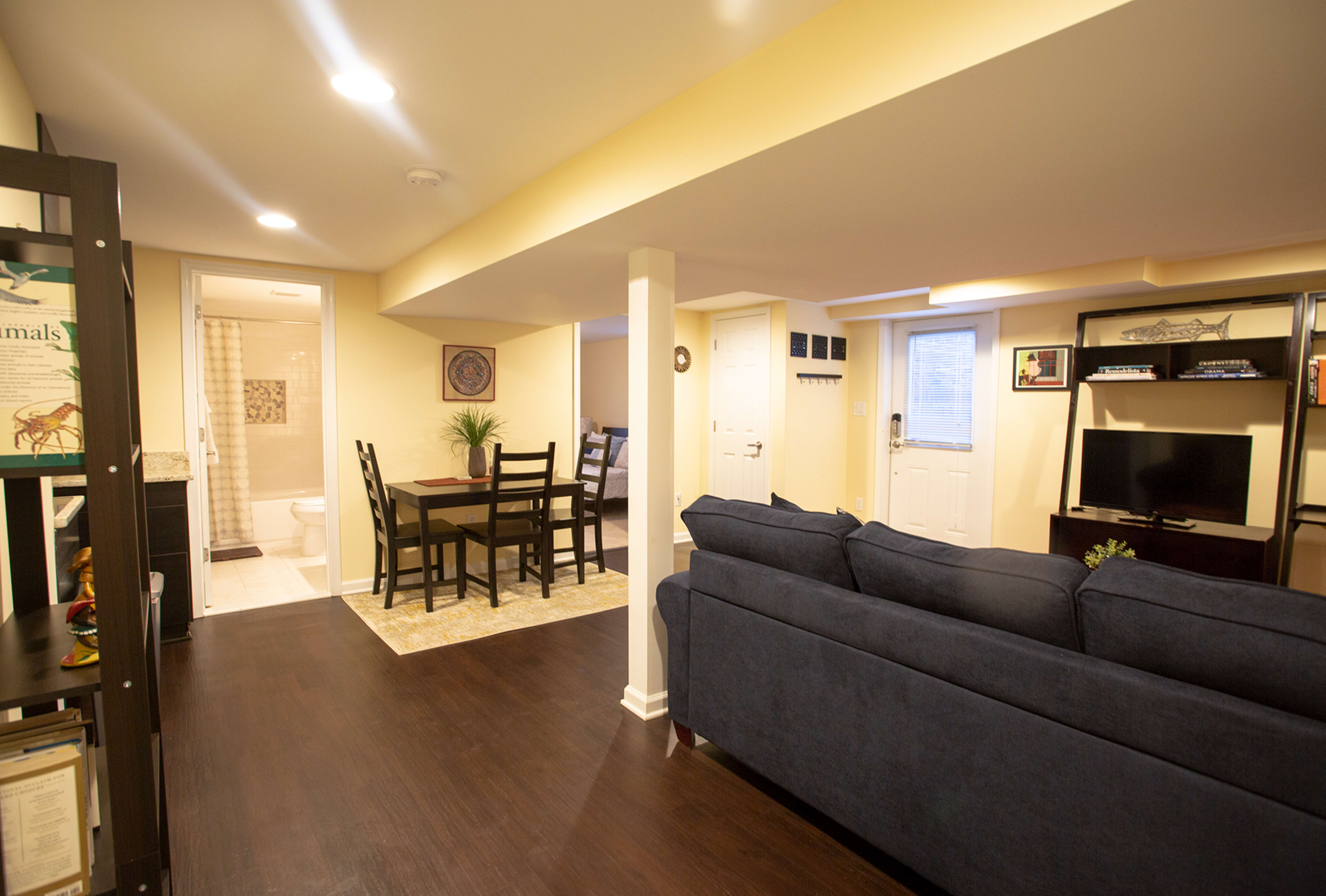 The image shows a finished basement with a seating area featuring a dark blue couch, a dining table with chairs, and a small kitchen area leading to a bathroom and an entrance door.