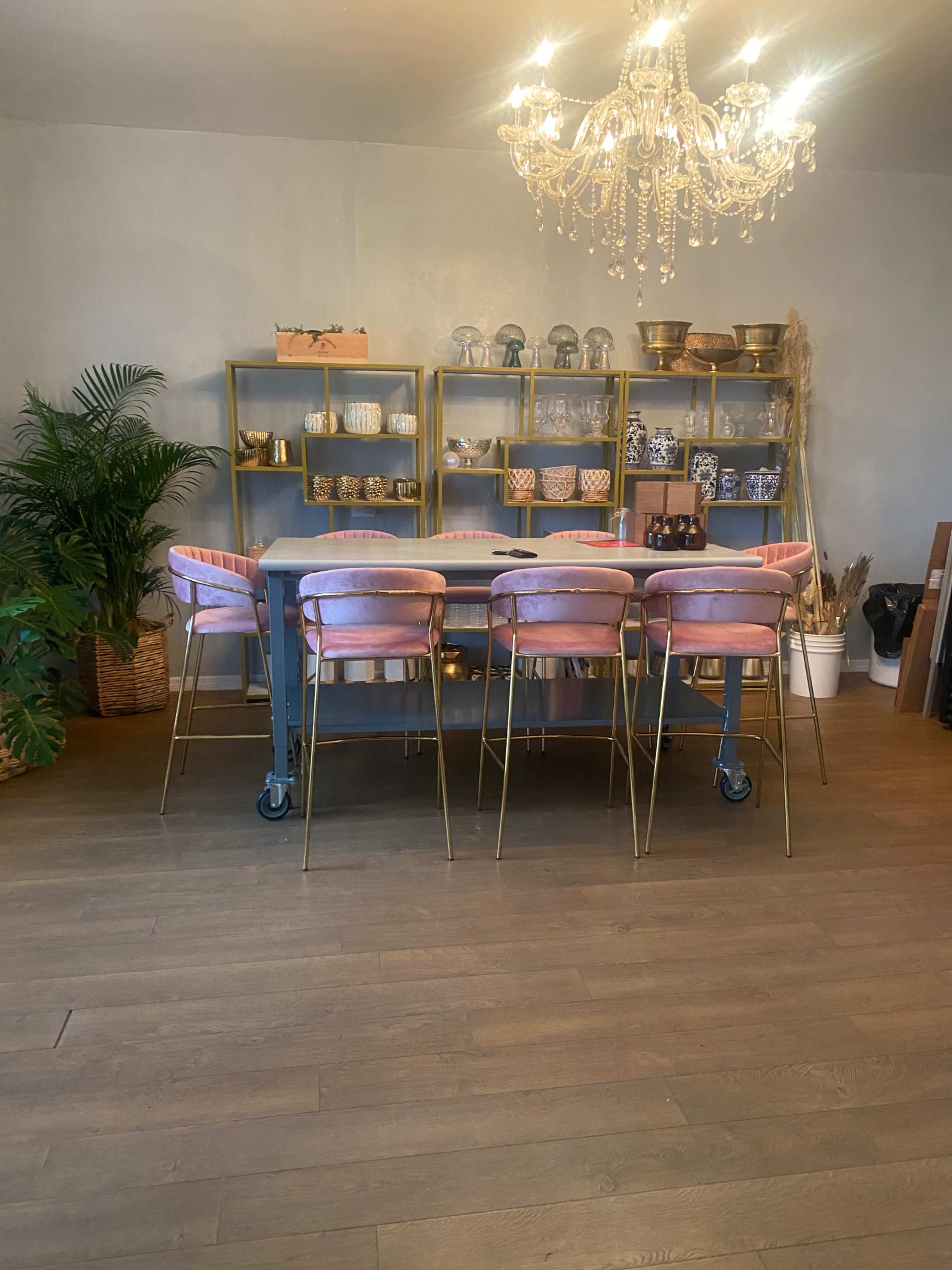 A dining area features a gray table surrounded by pink upholstered chairs on wheels and a gold chandelier above, with neatly organized shelves displaying various decorative items.