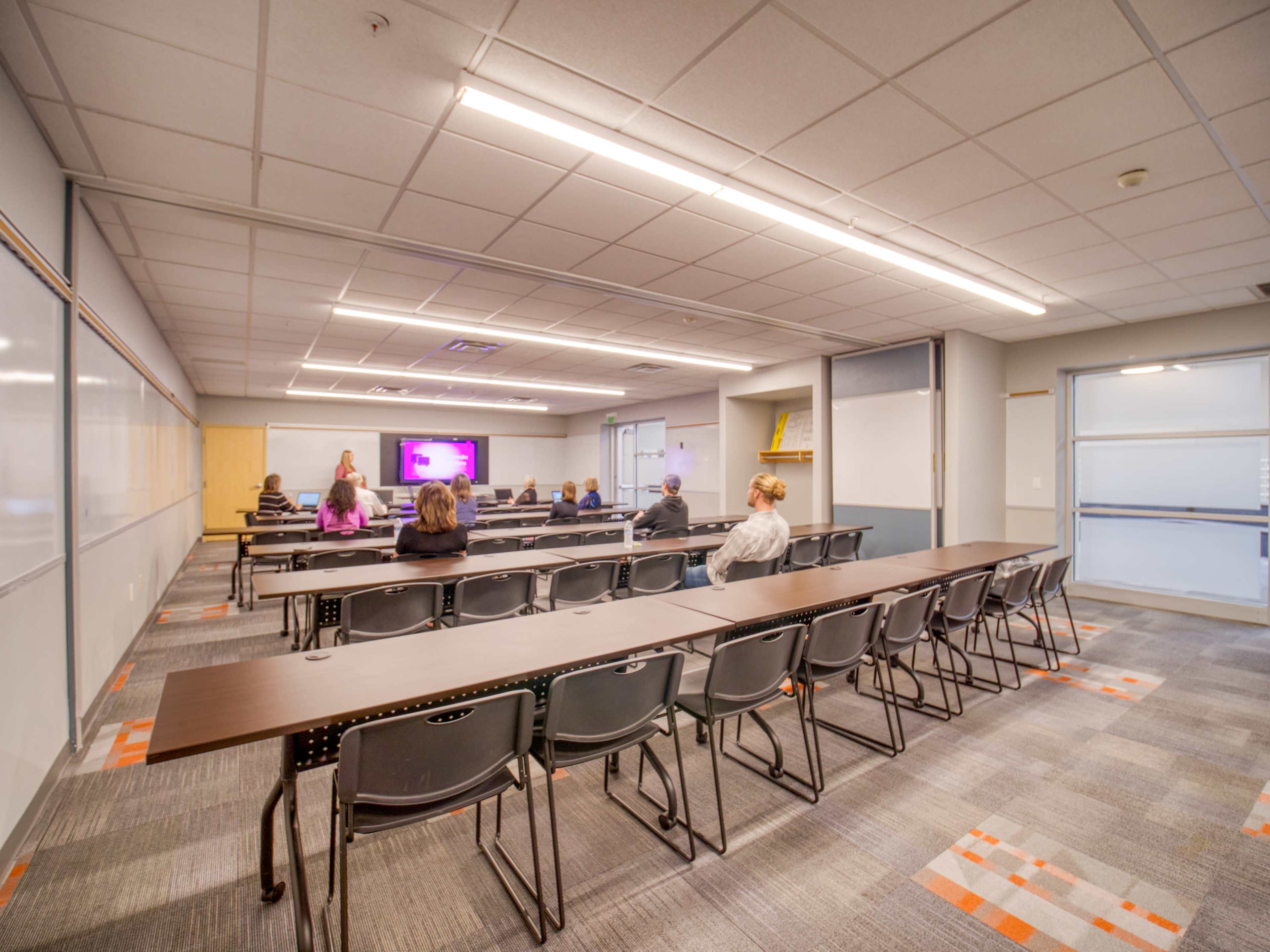 A classroom features rows of tables and chairs arranged facing a screen, with a few individuals seated and a presenter near the front.