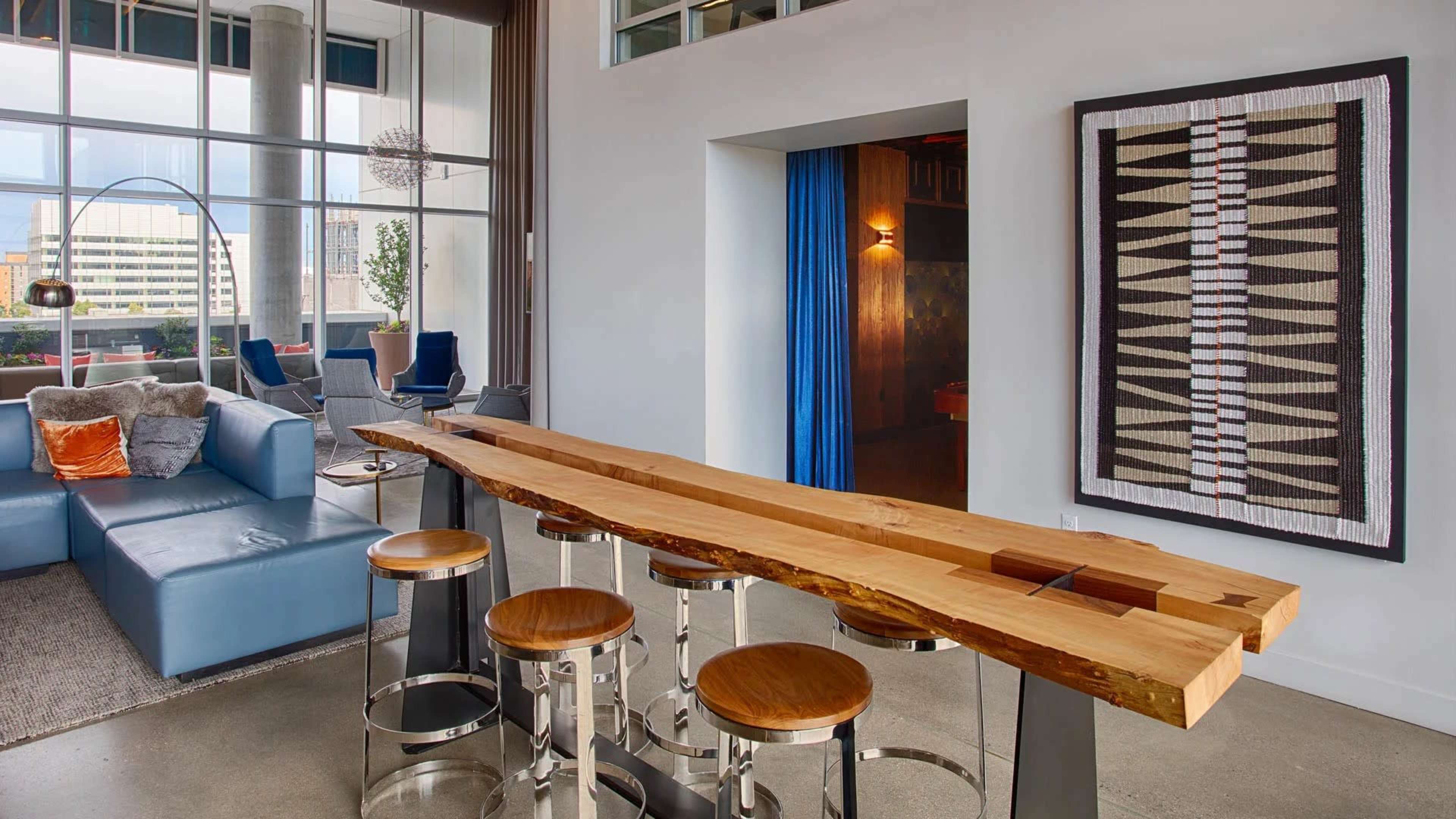 A modern interior with a sleek wooden bar table and four stools, situated next to a seating area and a doorway leading to another room.