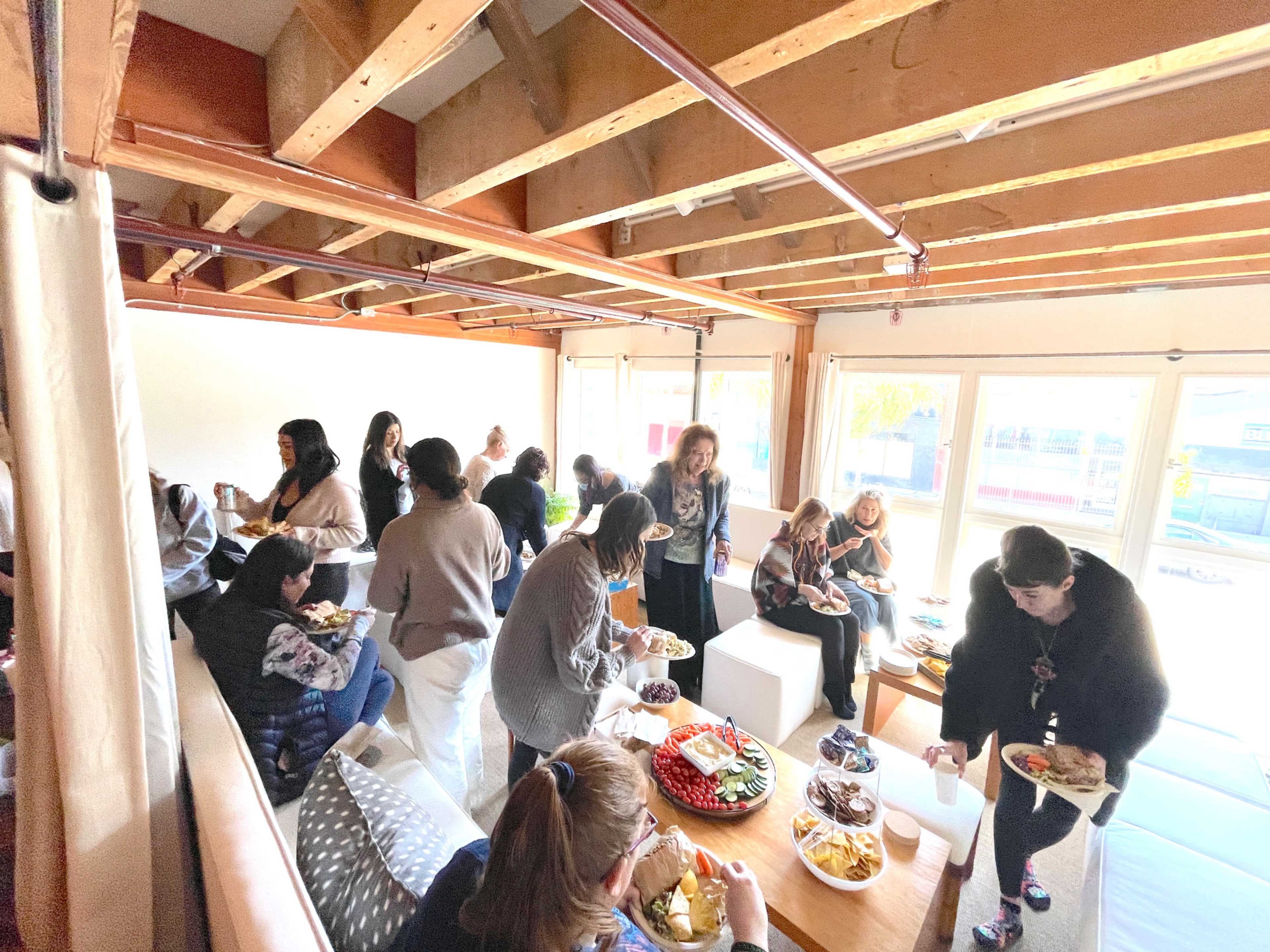 A group of people is gathered in a room with exposed beams, enjoying a buffet-style meal while socializing.