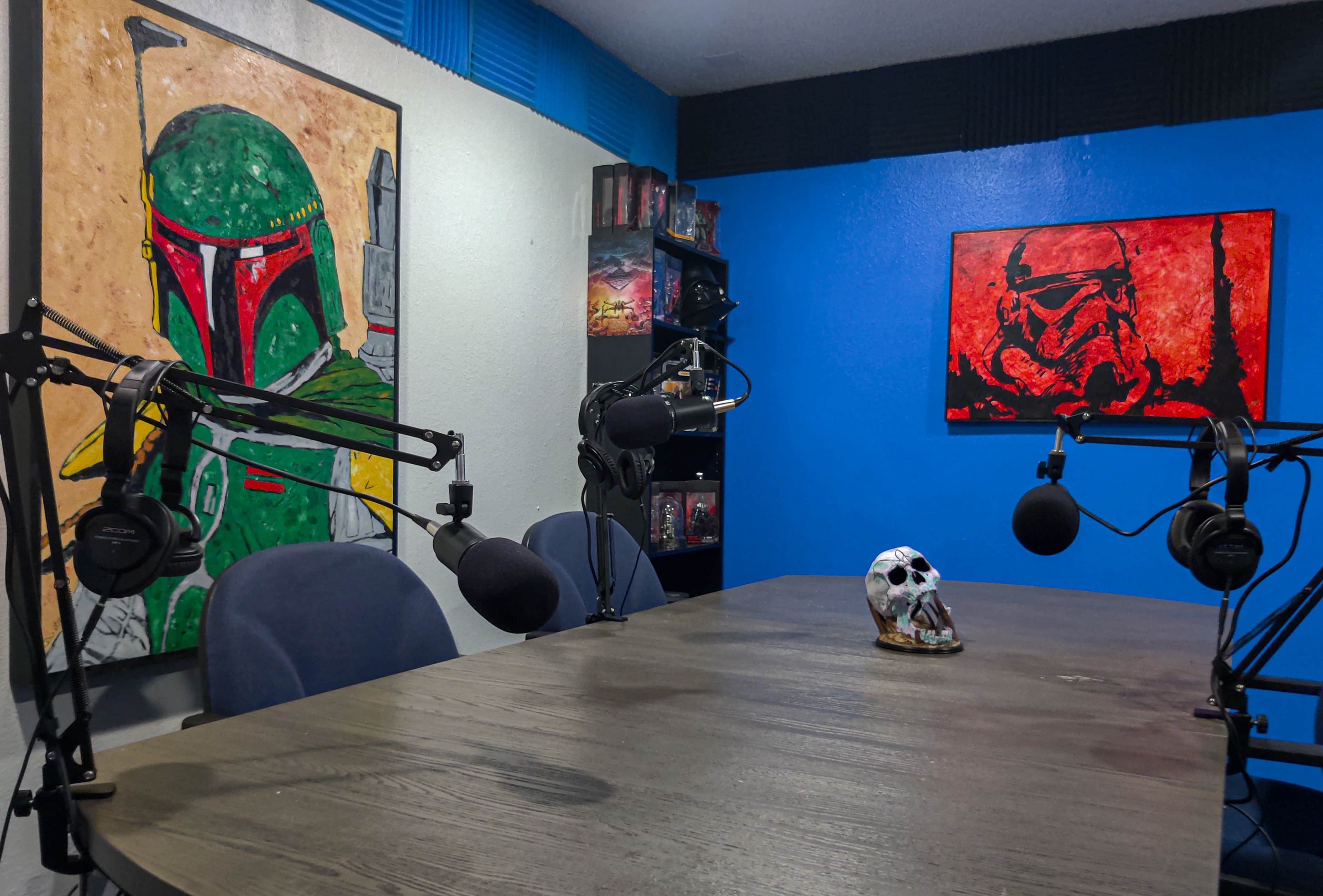 A podcast studio with a wooden table, two microphone setups, and artwork featuring Boba Fett and a skull figurine on the table.