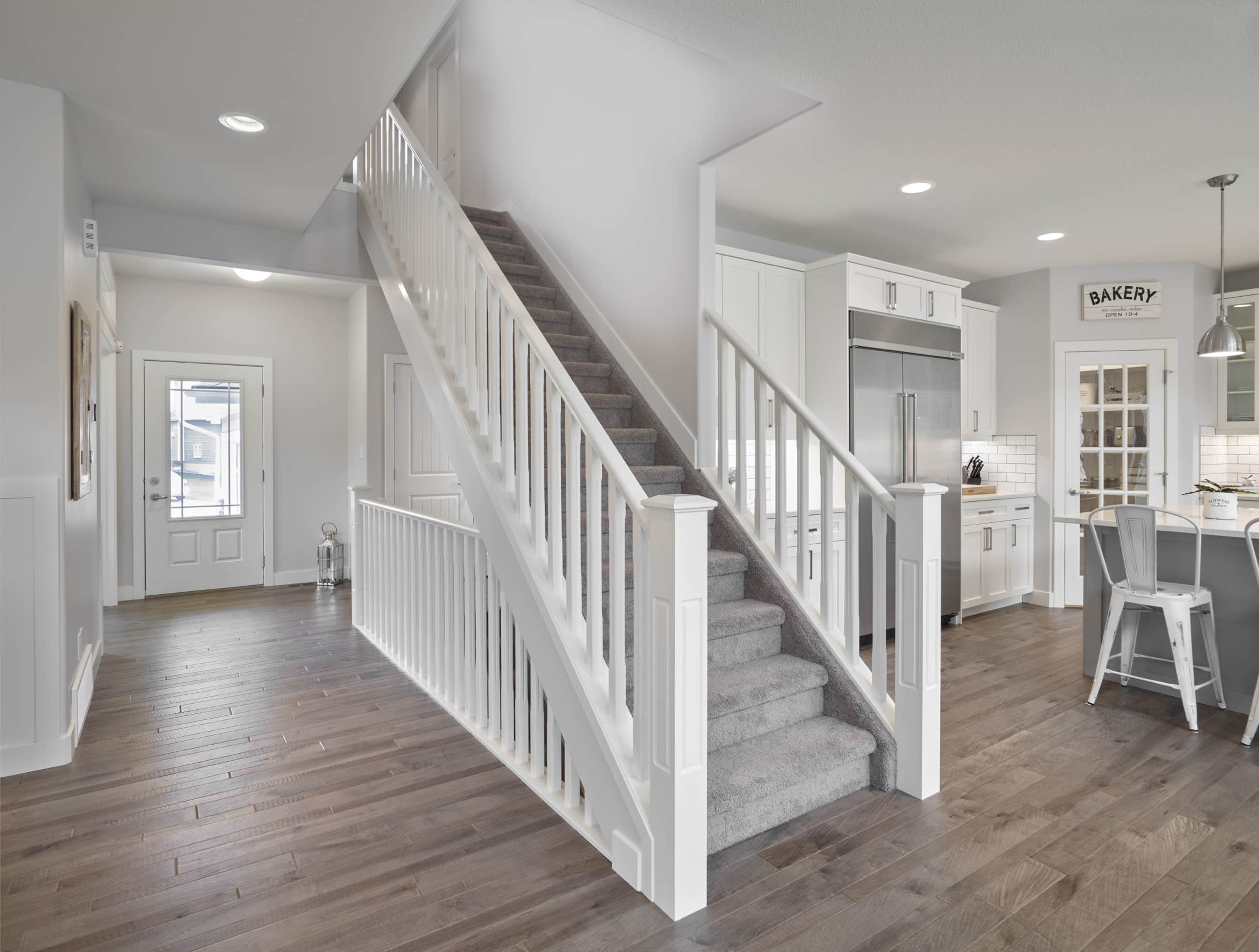 A well-lit modern interior features a staircase leading to an upper level, with hardwood floors and a kitchen area visible on the right.