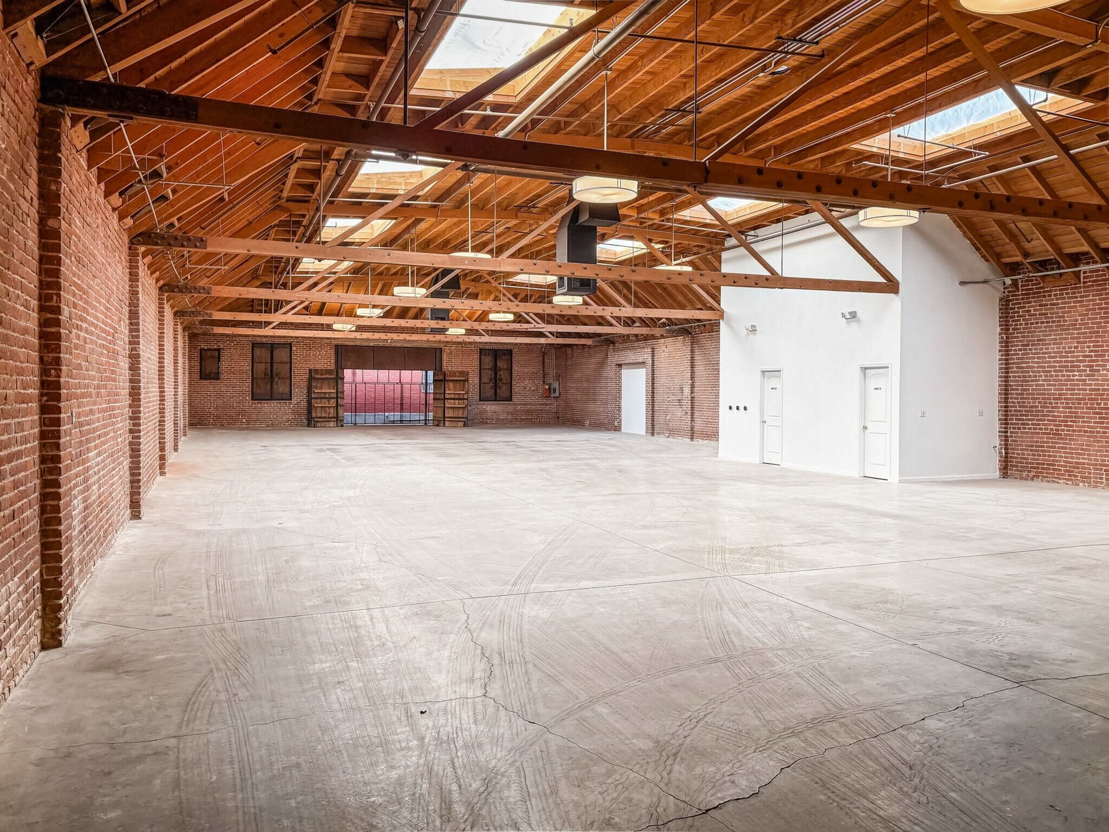 The image shows a spacious interior of a building with exposed brick walls, wooden beams, and a concrete floor, featuring two doors on one side.