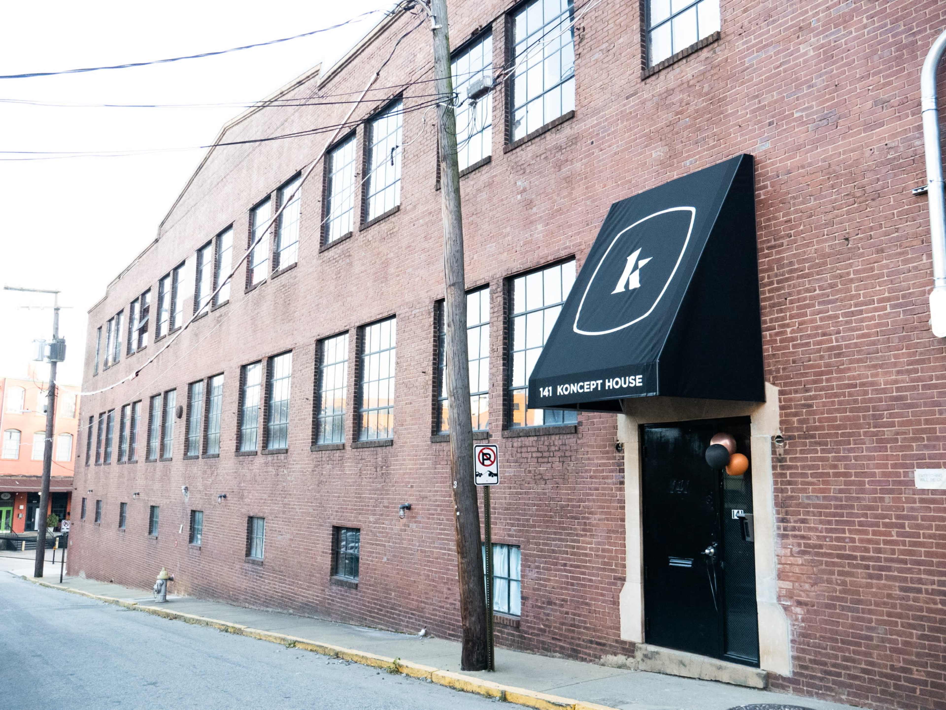 The image shows a brick building with large windows and a black awning featuring the letter "K" above the entrance labeled "KONCEPT HOUSE."