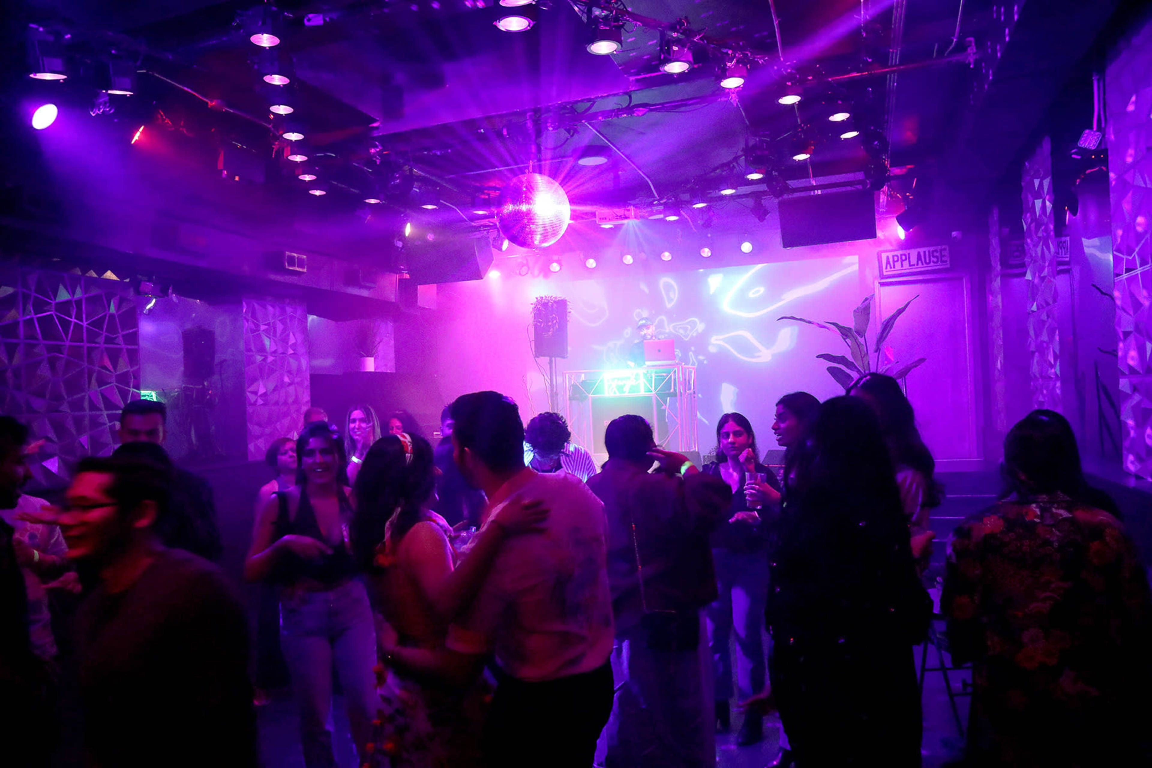 A lively crowd dances under colorful lights in a nightclub featuring a disco ball and a DJ setup.