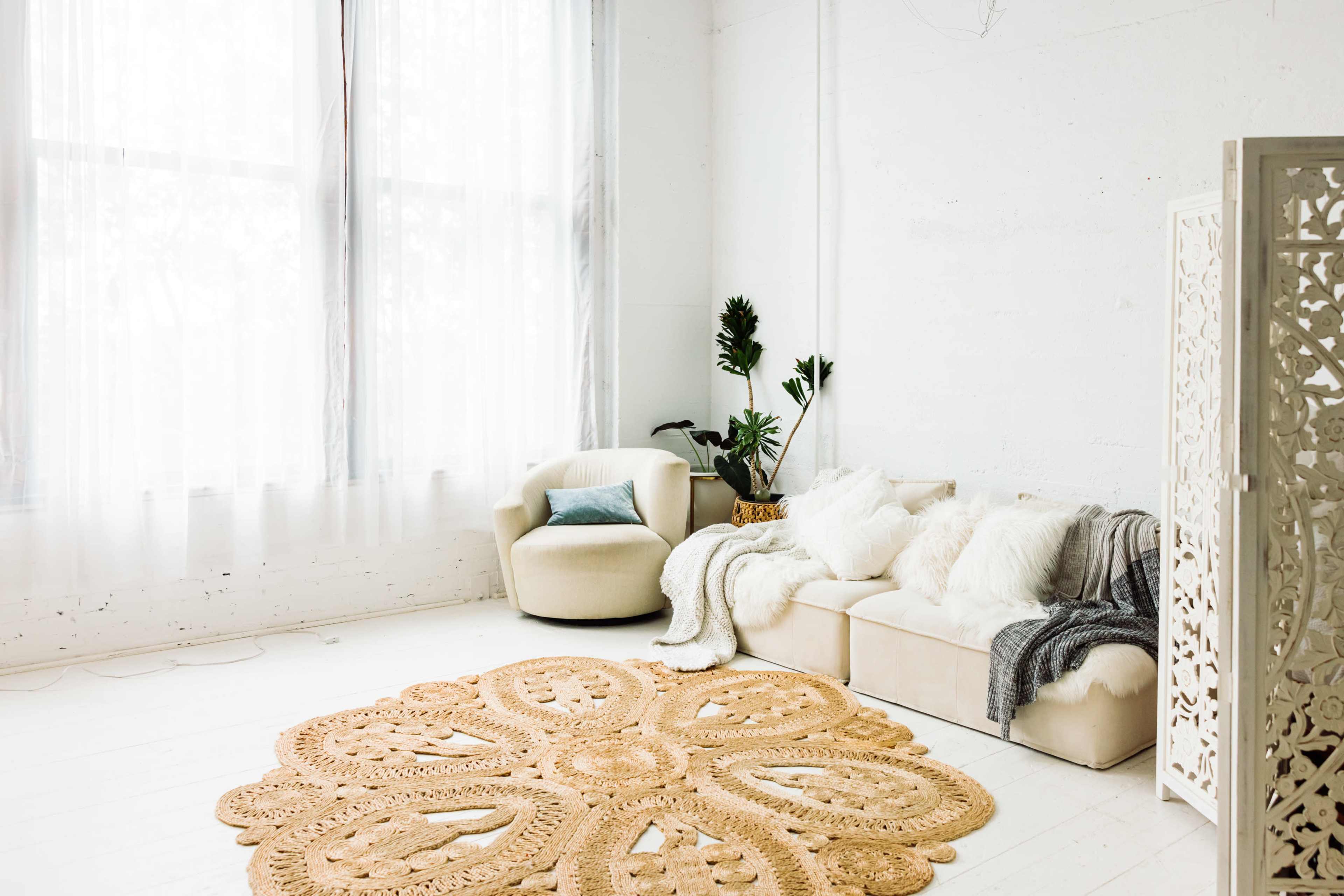 The image shows a bright, minimalist living space with a cream-colored armchair, a sofa draped with soft blankets, and a large round woven rug on a light wooden floor.