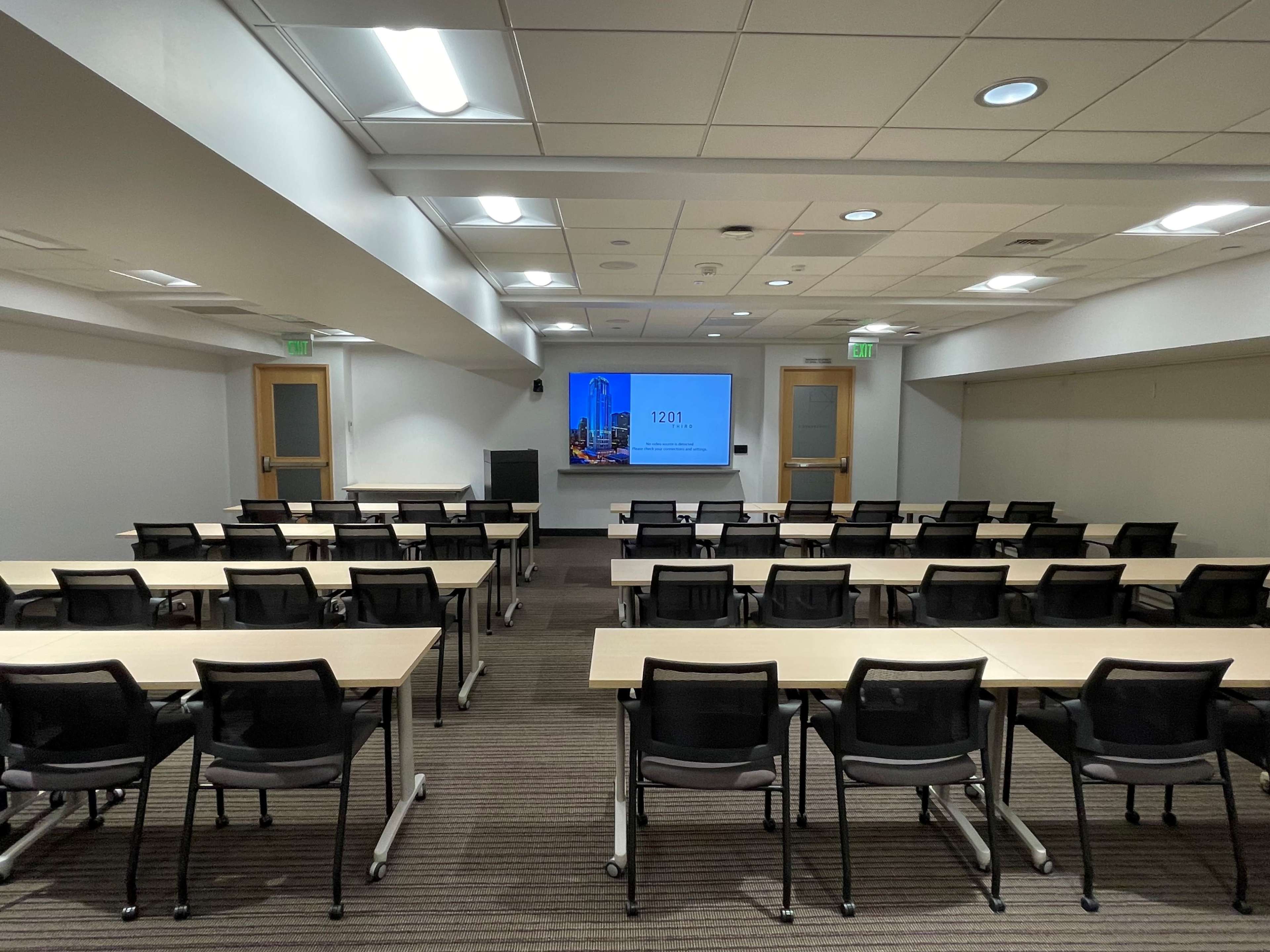 A conference room with rows of tables and chairs facing a screen displaying a presentation, illuminated by overhead lighting.
