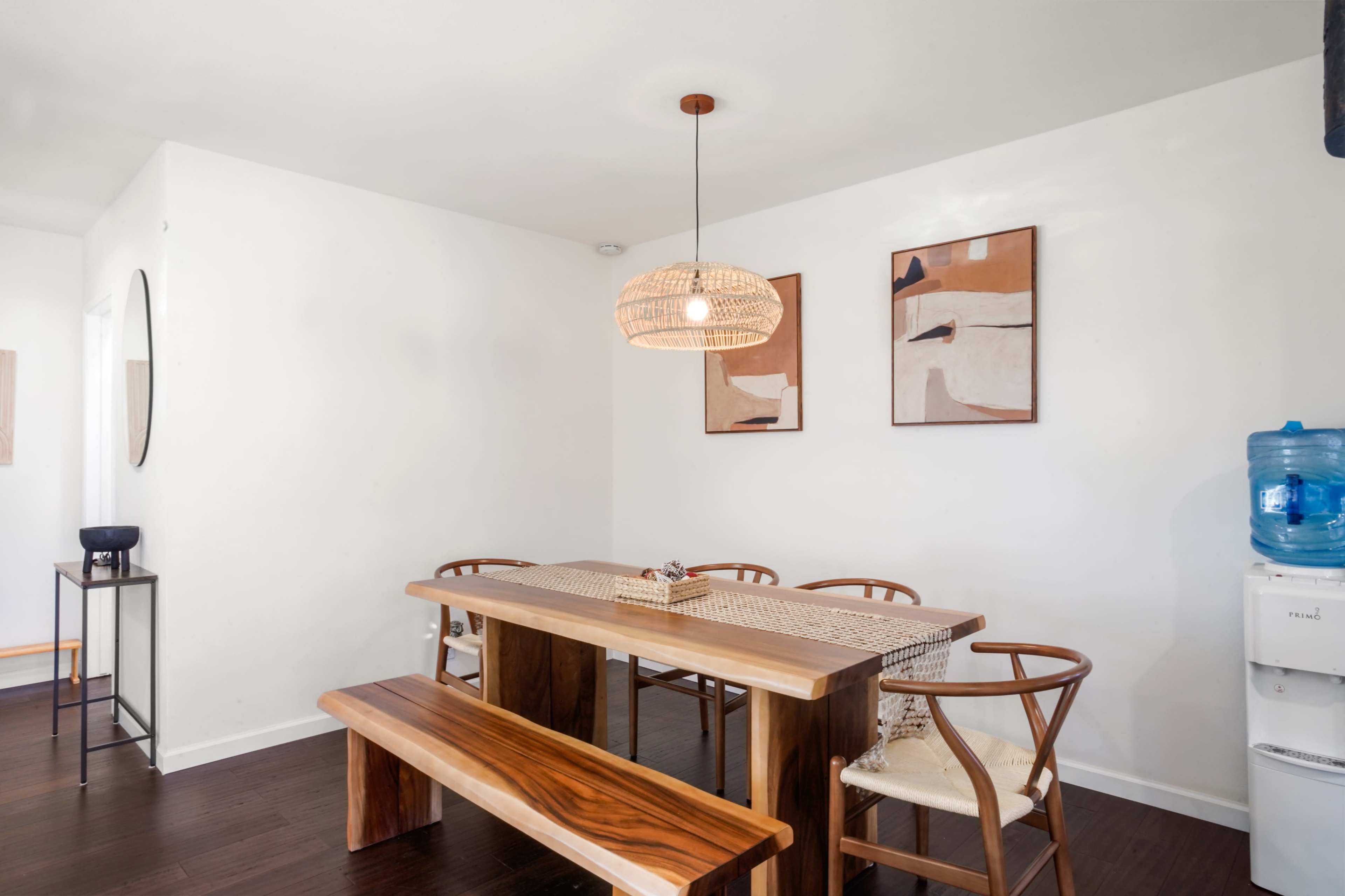 A simple dining area features a wooden table and bench, accompanied by stylish chairs, with a pendant light overhead and wall art displayed.