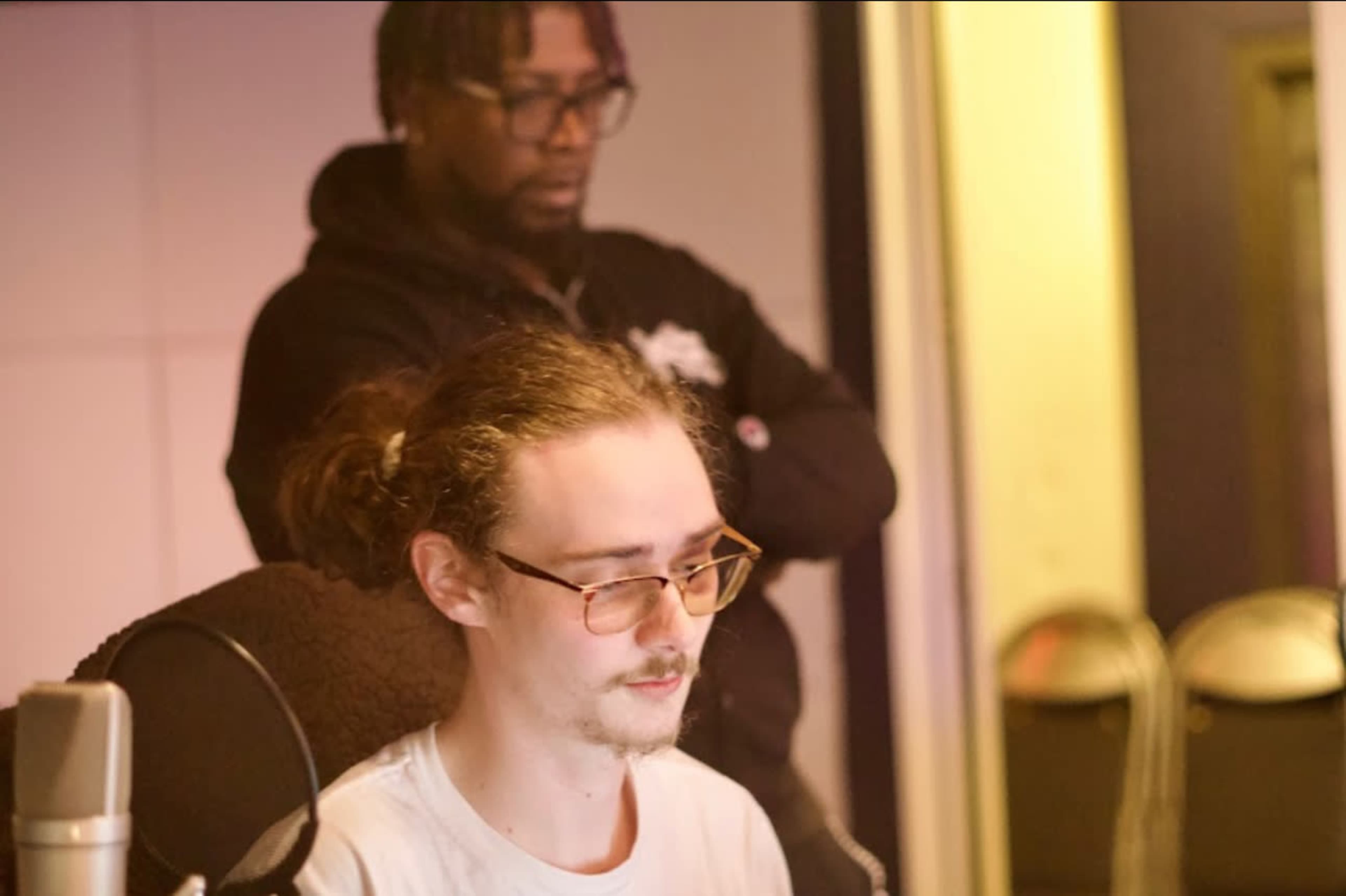A person with glasses sits at a recording console while another person stands behind them in a studio setting.