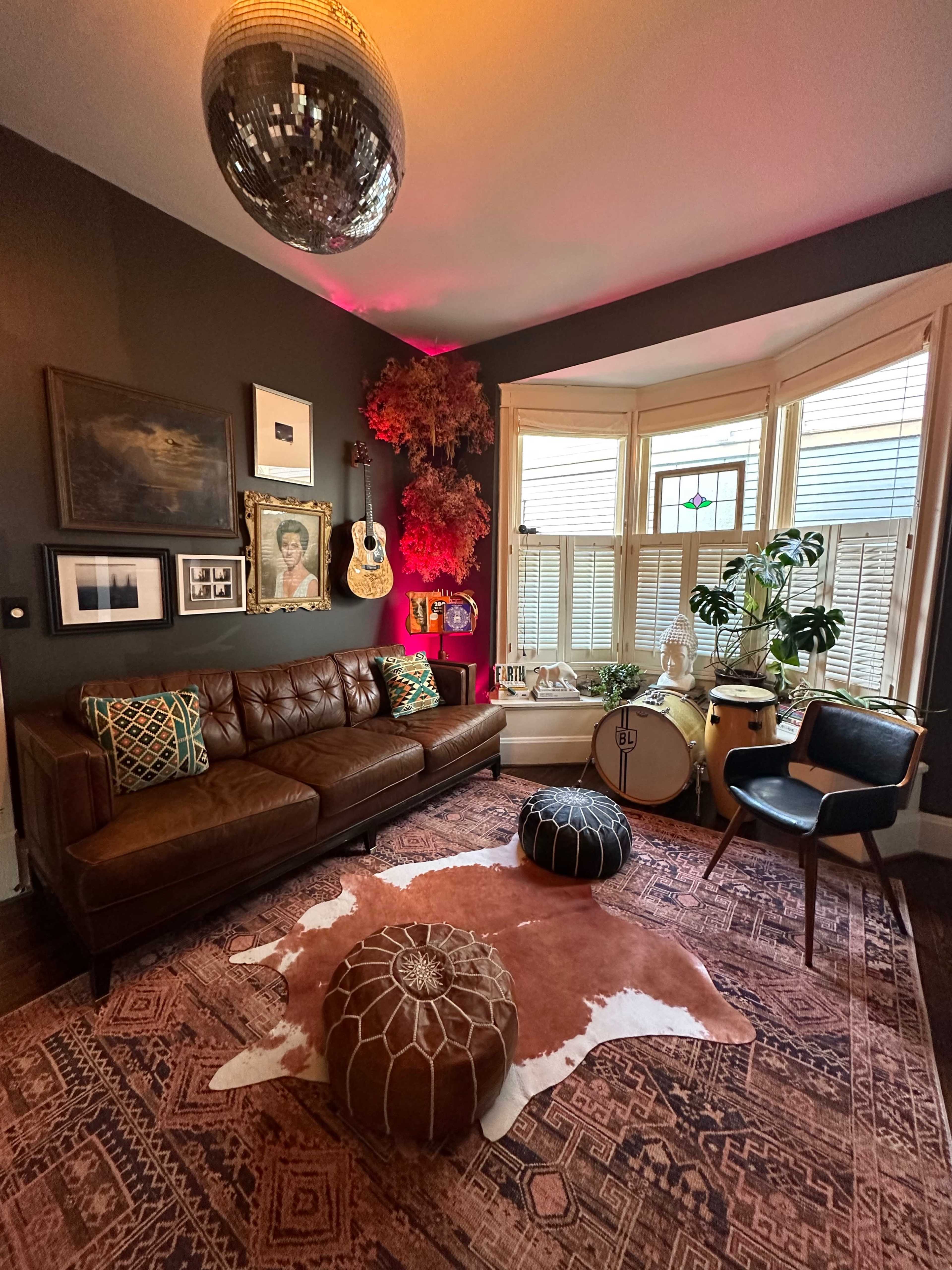 A cozy living room features a brown leather sofa, decorative poufs, a patterned rug, and a bay window filled with plants and art.