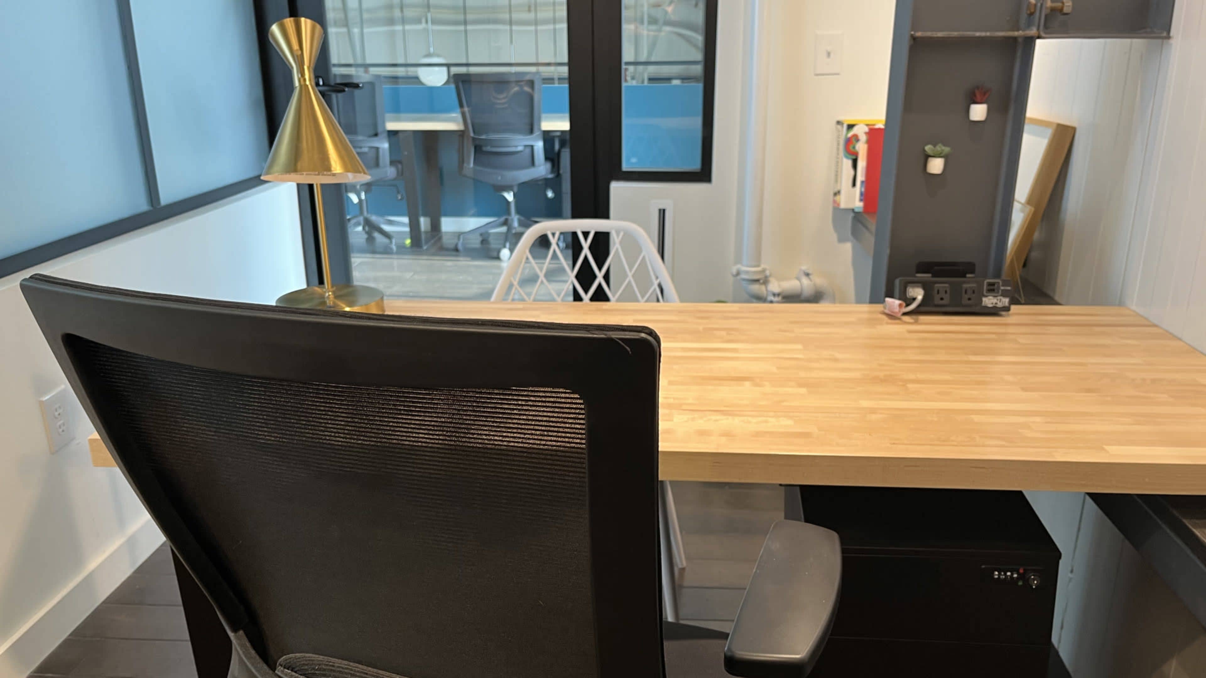 A modern desk with a wooden tabletop, an office chair, a gold lamp, and a view of an office space through a large window.