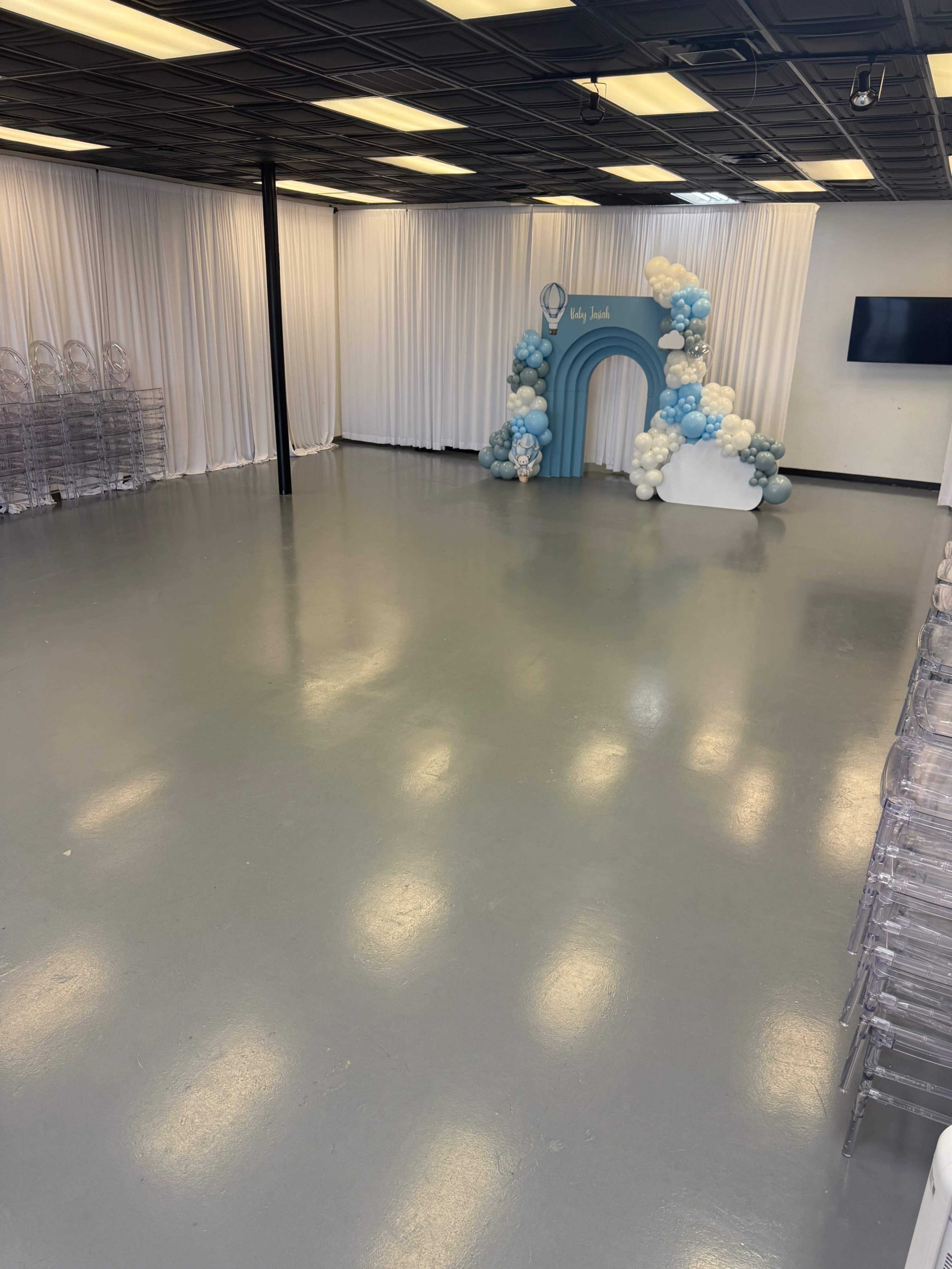 The image shows a spacious event room with a light gray floor, white draped walls, a balloon arch in blue and white, and clear acrylic chairs lined up against one wall.