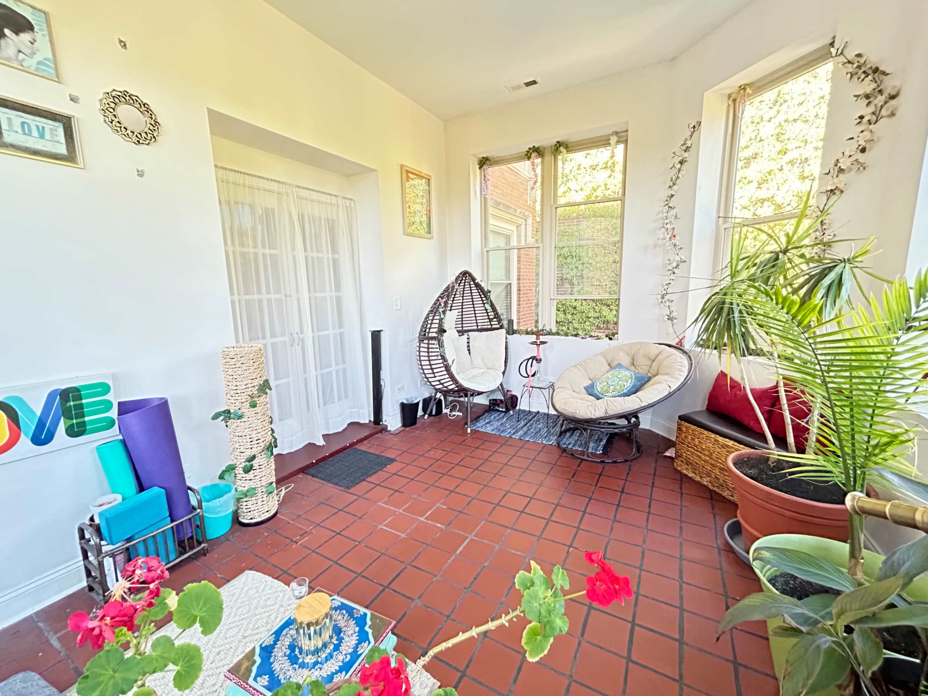 The image shows a bright sunroom with a hanging chair, cushioned seating, plants, and decorative elements on the walls.