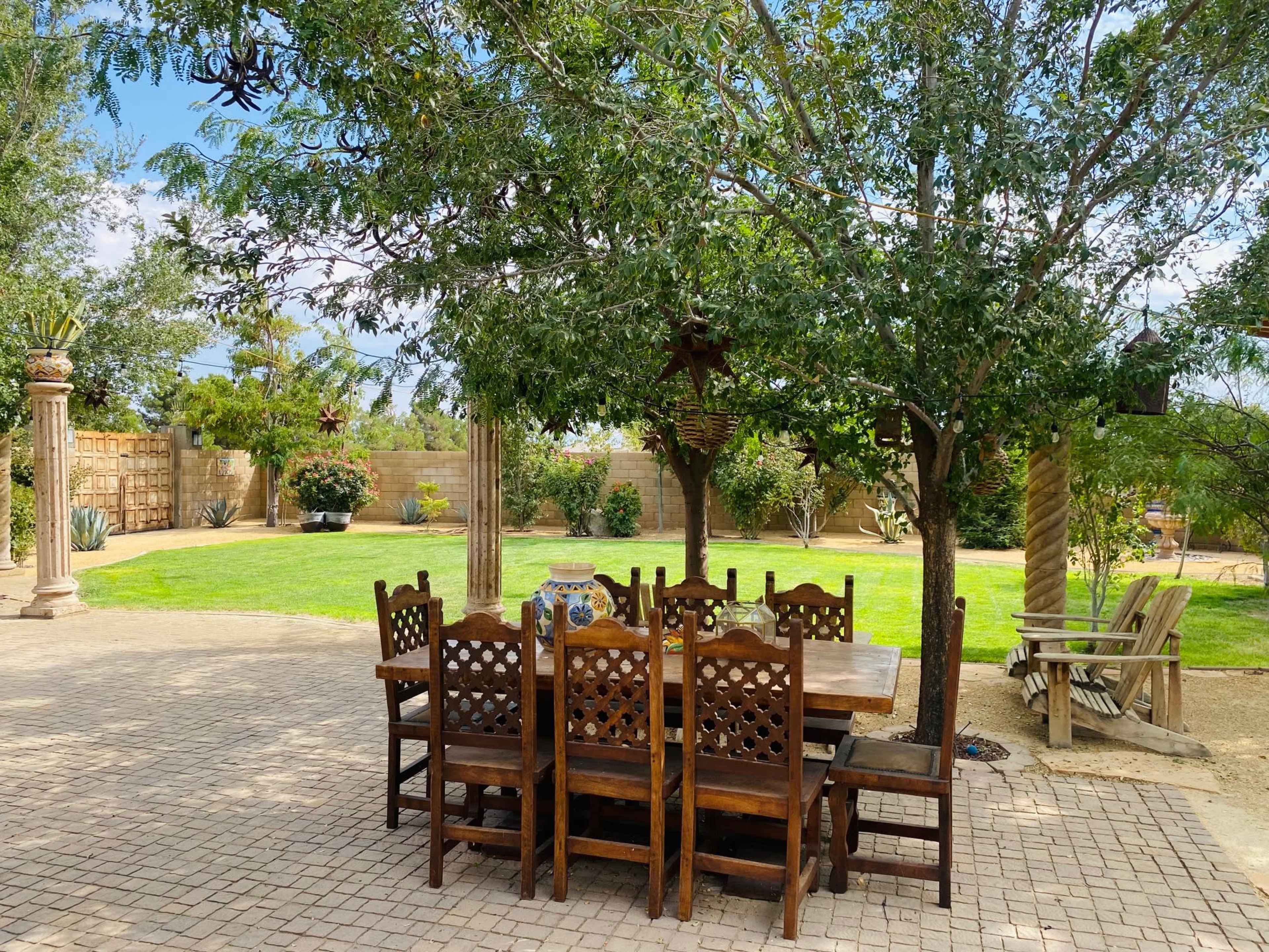 A wooden dining table surrounded by several chairs sits under a large tree in a grassy outdoor area.