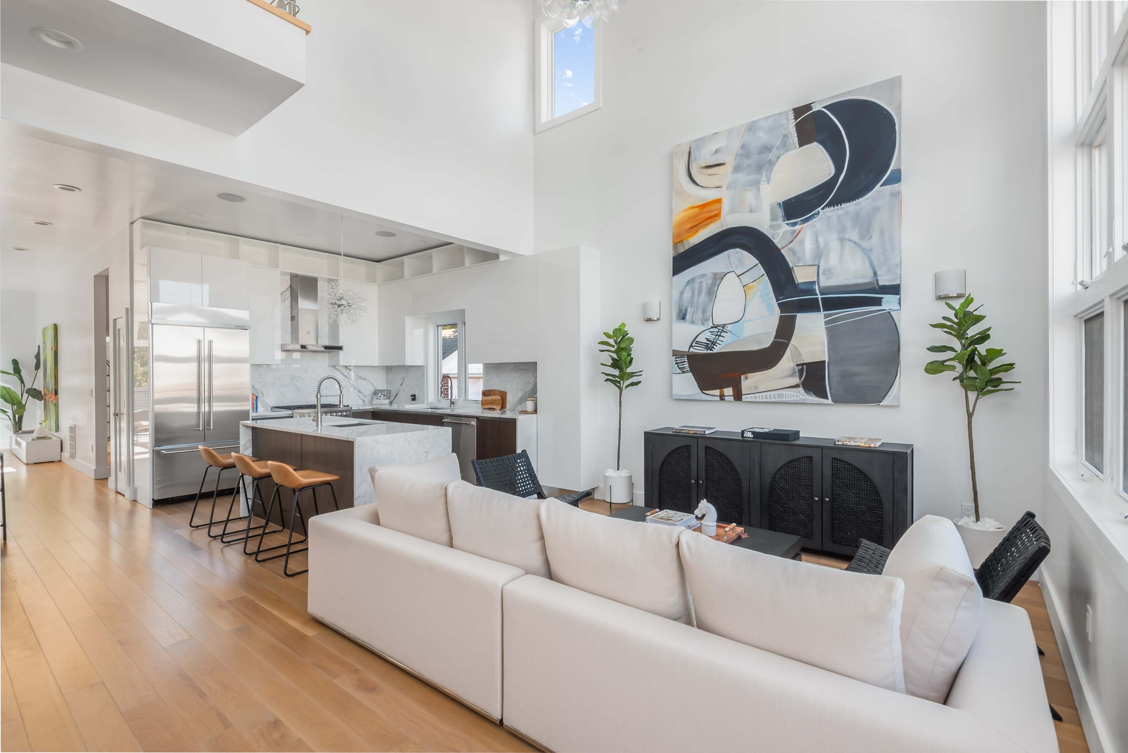 A modern living space with a white sofa, a kitchen area featuring stainless steel appliances, and a large abstract painting on the wall.