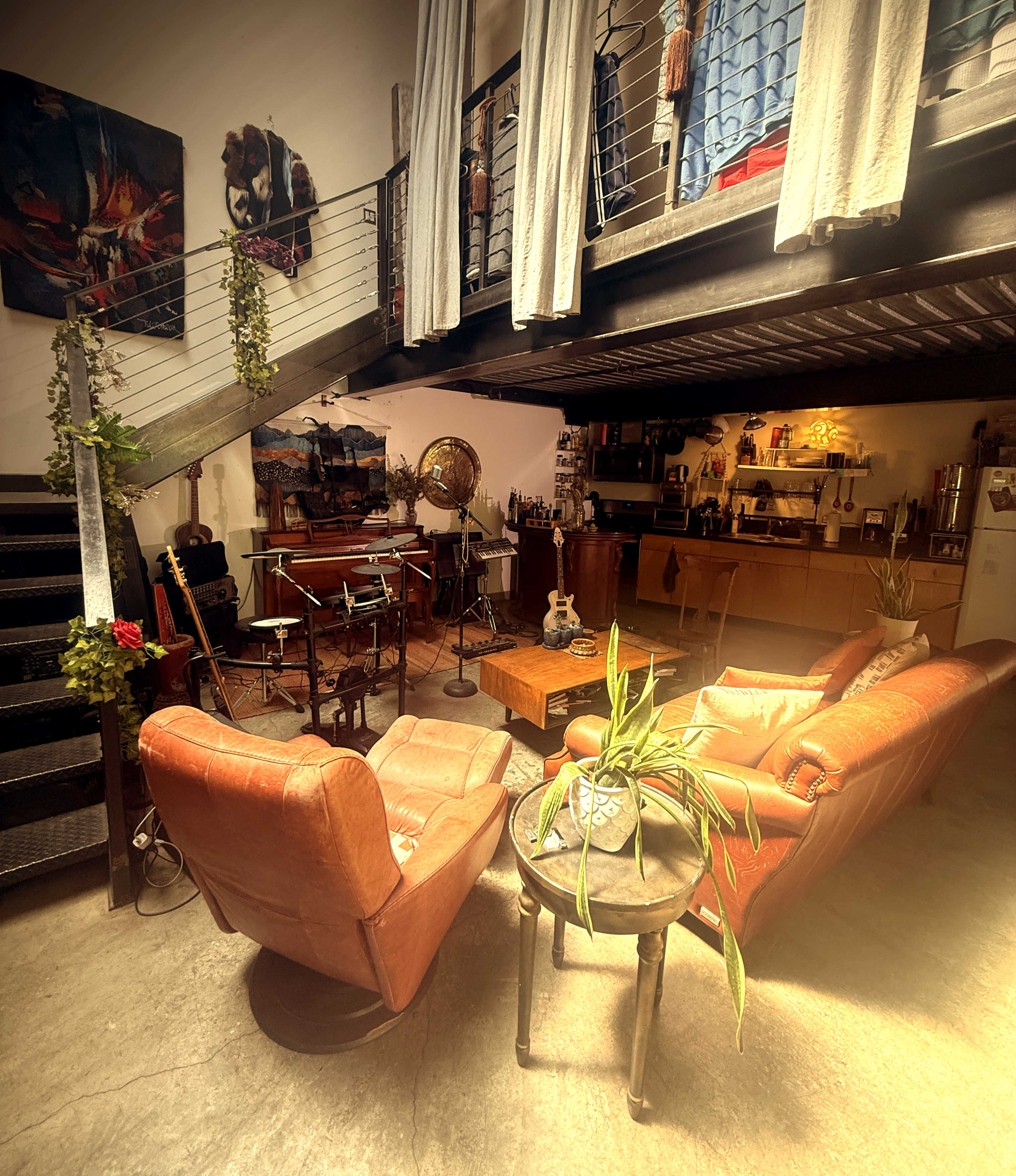 The image shows a cozy urban living space with leather armchairs, a wooden coffee table, and an upper balcony overlooking a kitchen area.