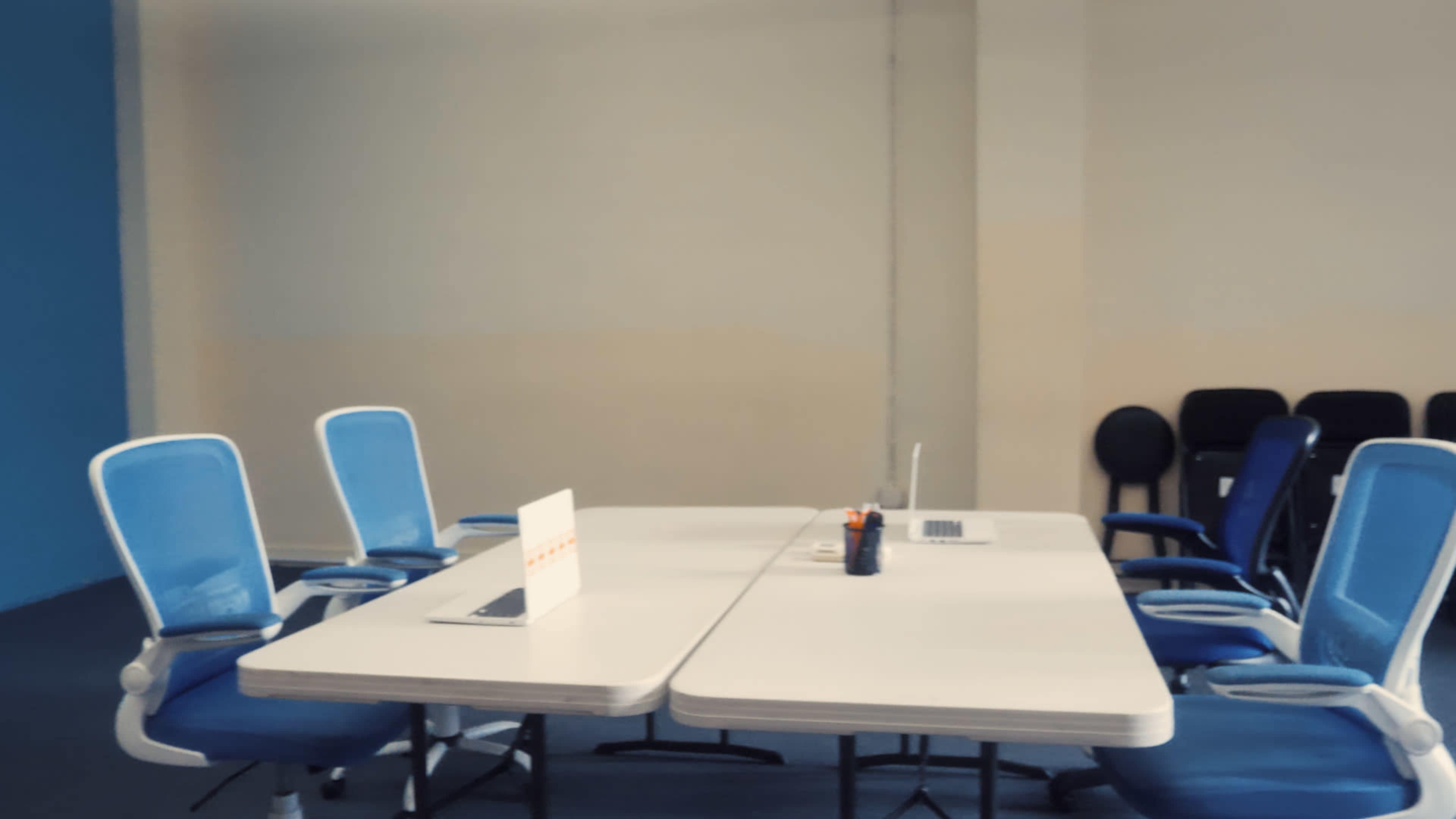 A conference room features two joining tables surrounded by blue and black chairs, with a laptop and a small sign on the table.