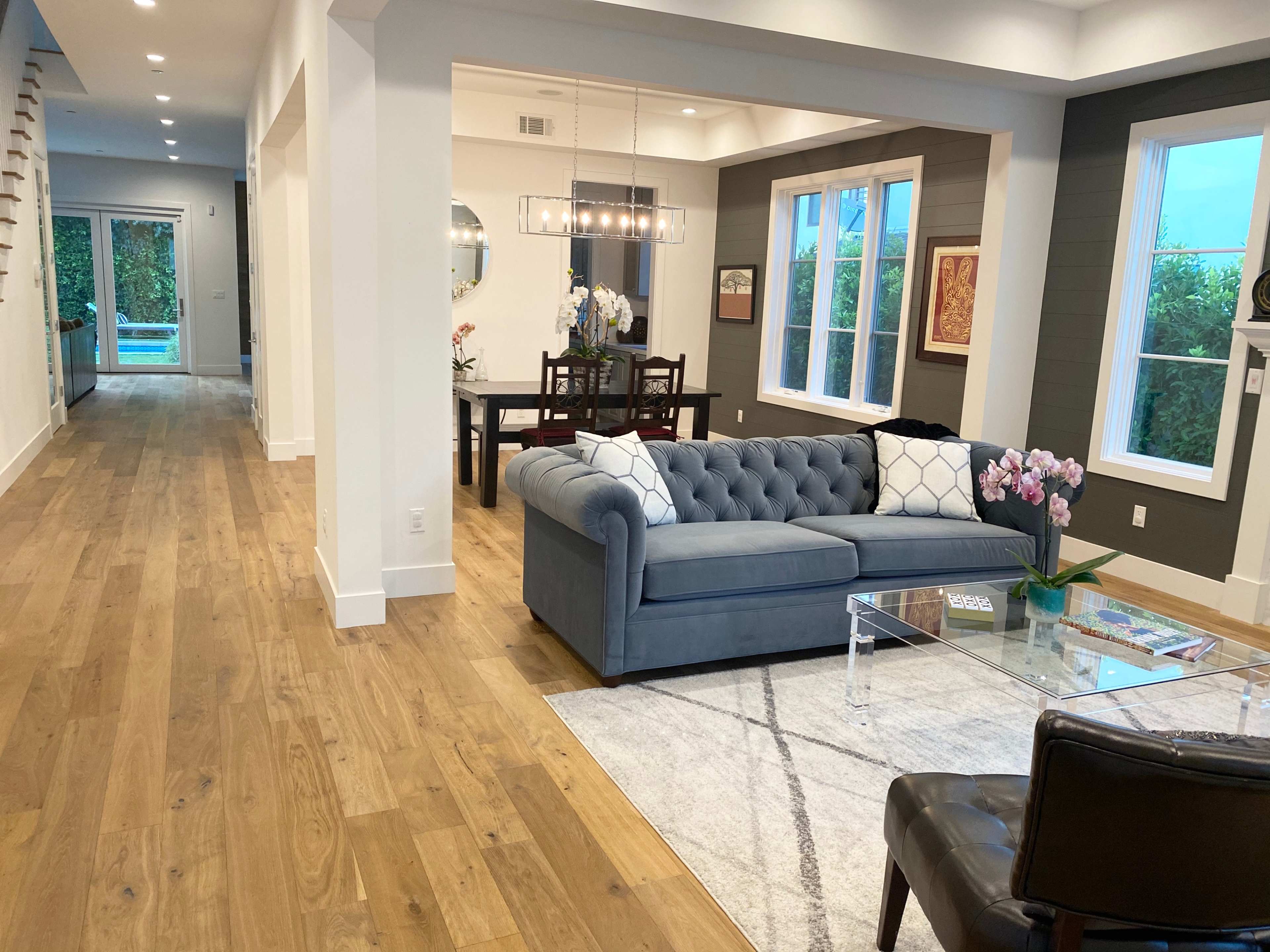 The image shows a modern living room with a gray upholstered sofa, a glass coffee table, and hardwood flooring, leading to a hallway with a dining area visible in the background.