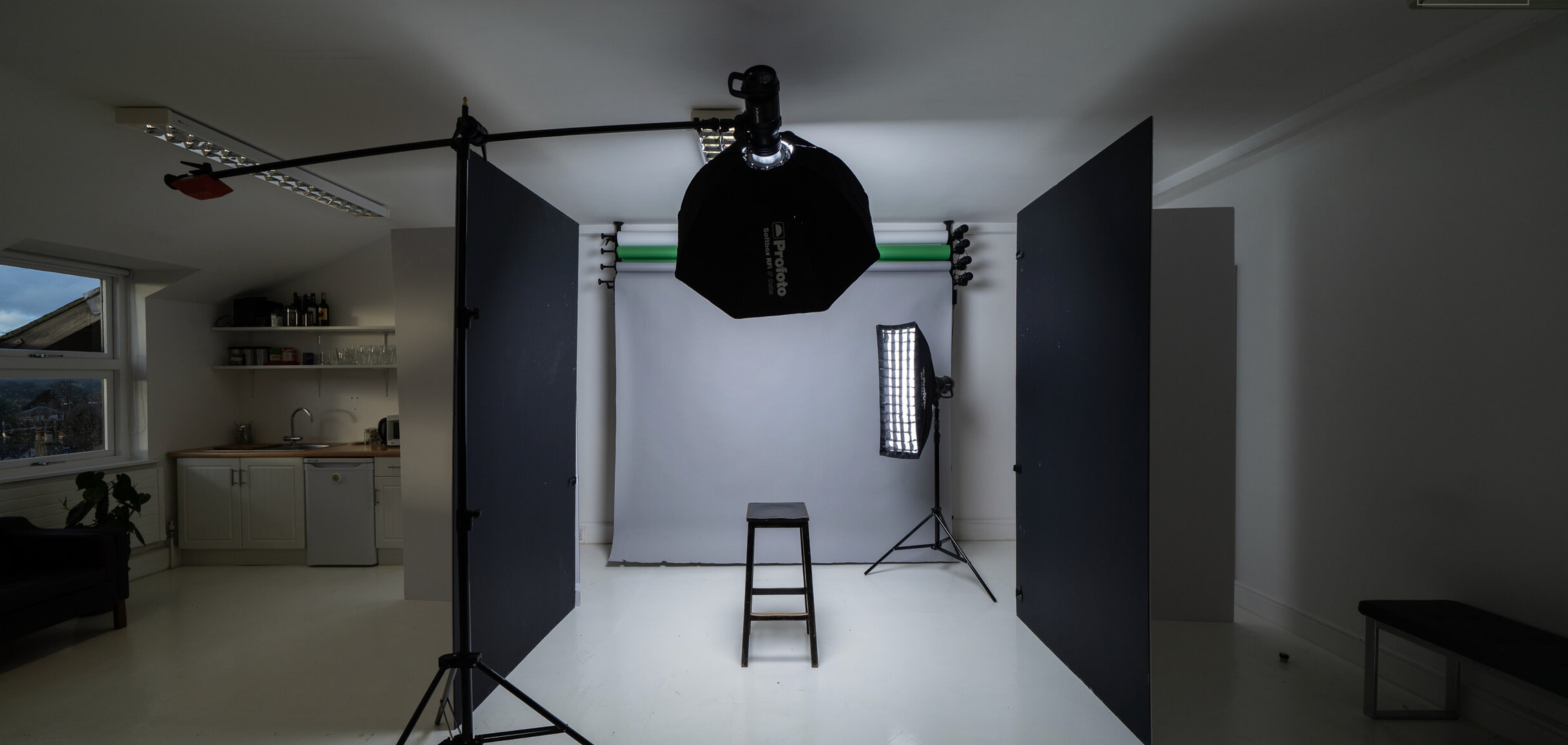 The image shows a photography studio with a stool placed in front of a gray backdrop, illuminated by a softbox and flanked by black panels.