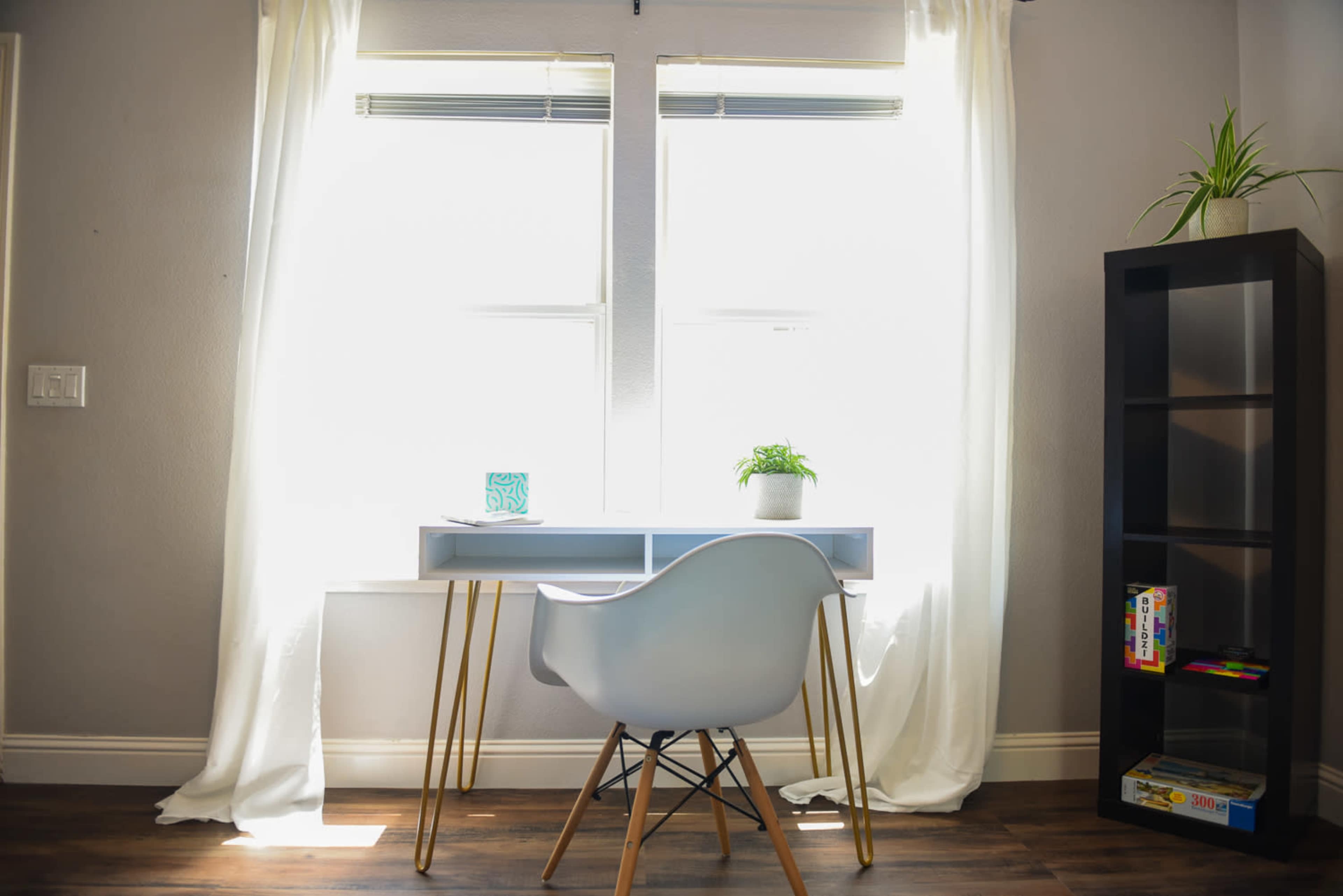 A modern desk with a chair and a potted plant is positioned beside two large windows covered by white curtains.