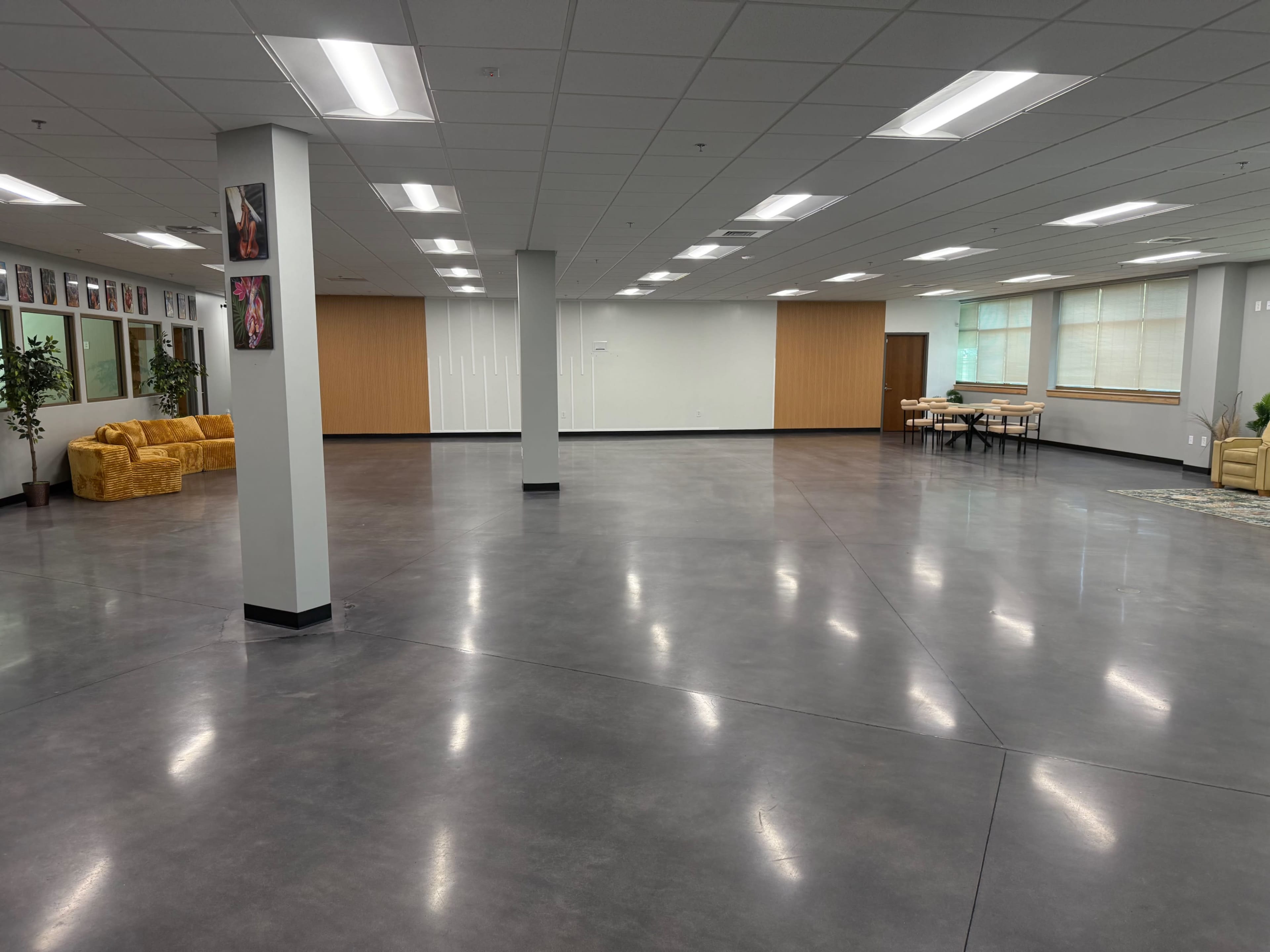 The image shows a spacious, empty room with polished concrete floors, white walls, and seating areas featuring yellow couches.