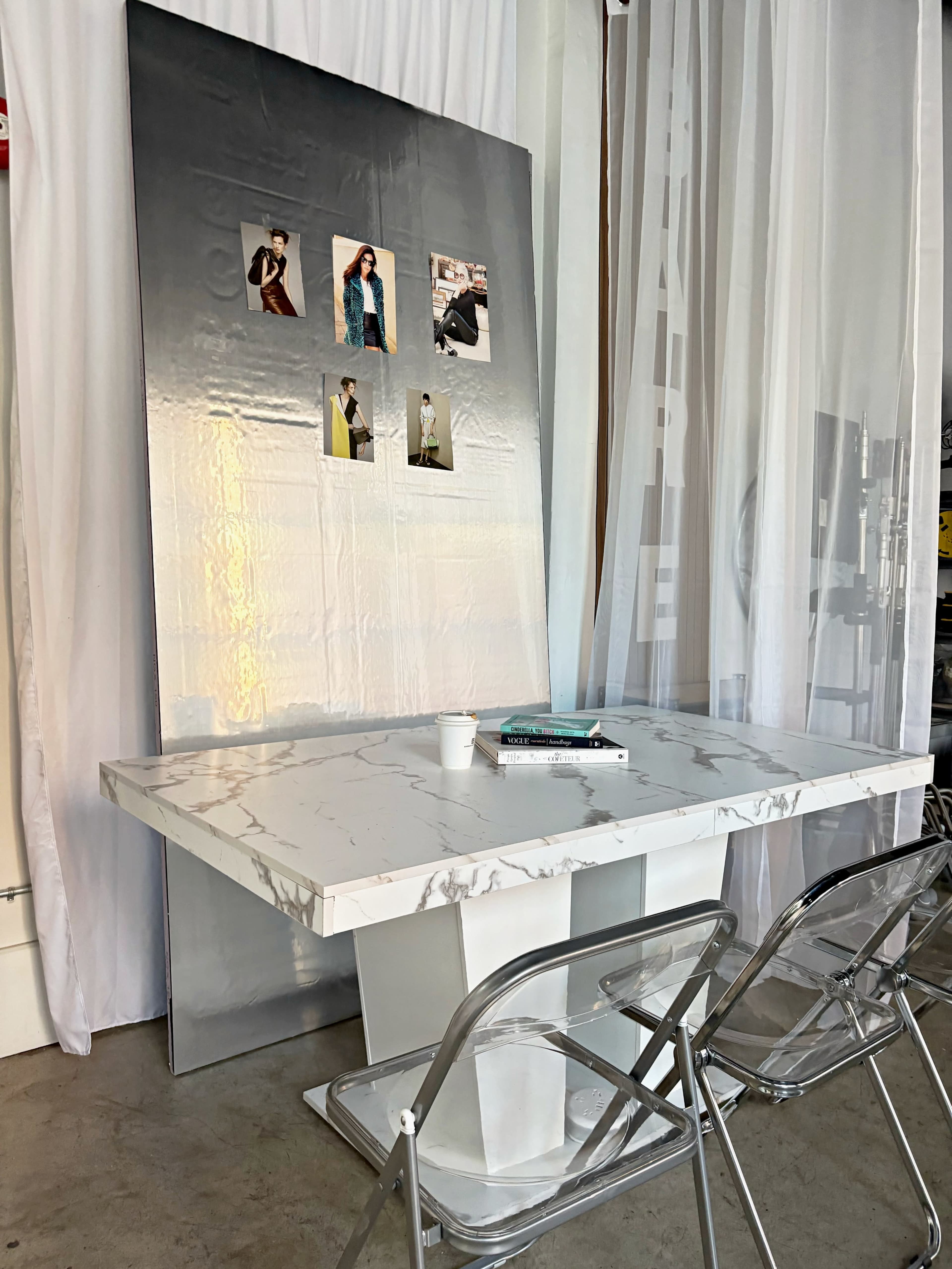 A modern table with a marble top and white base is set against a reflective silver backdrop, featuring four photos attached to it, with two transparent chairs nearby.