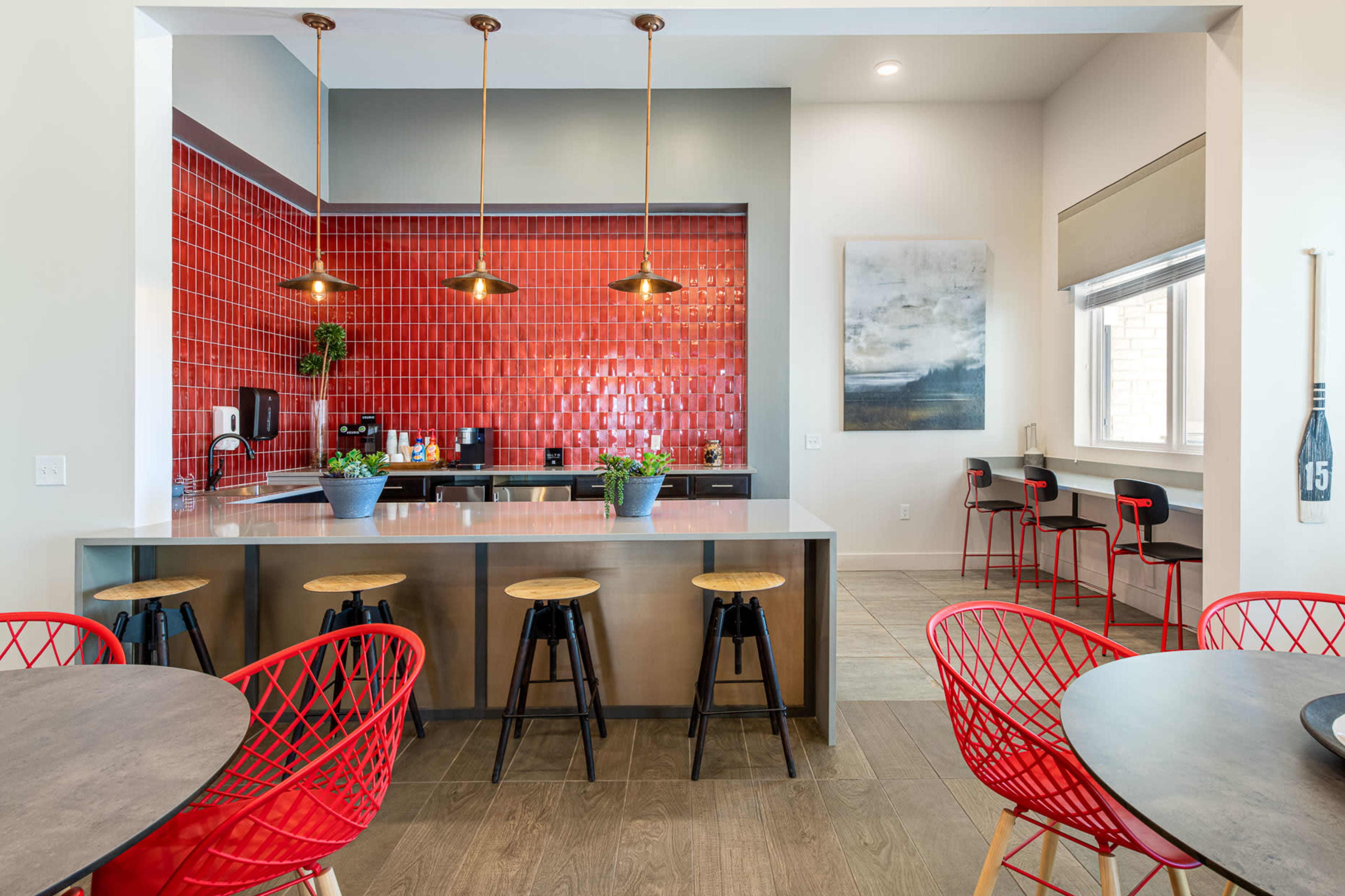 A modern kitchen featuring a red tiled backsplash, a central island with bar stools, and several dining tables.