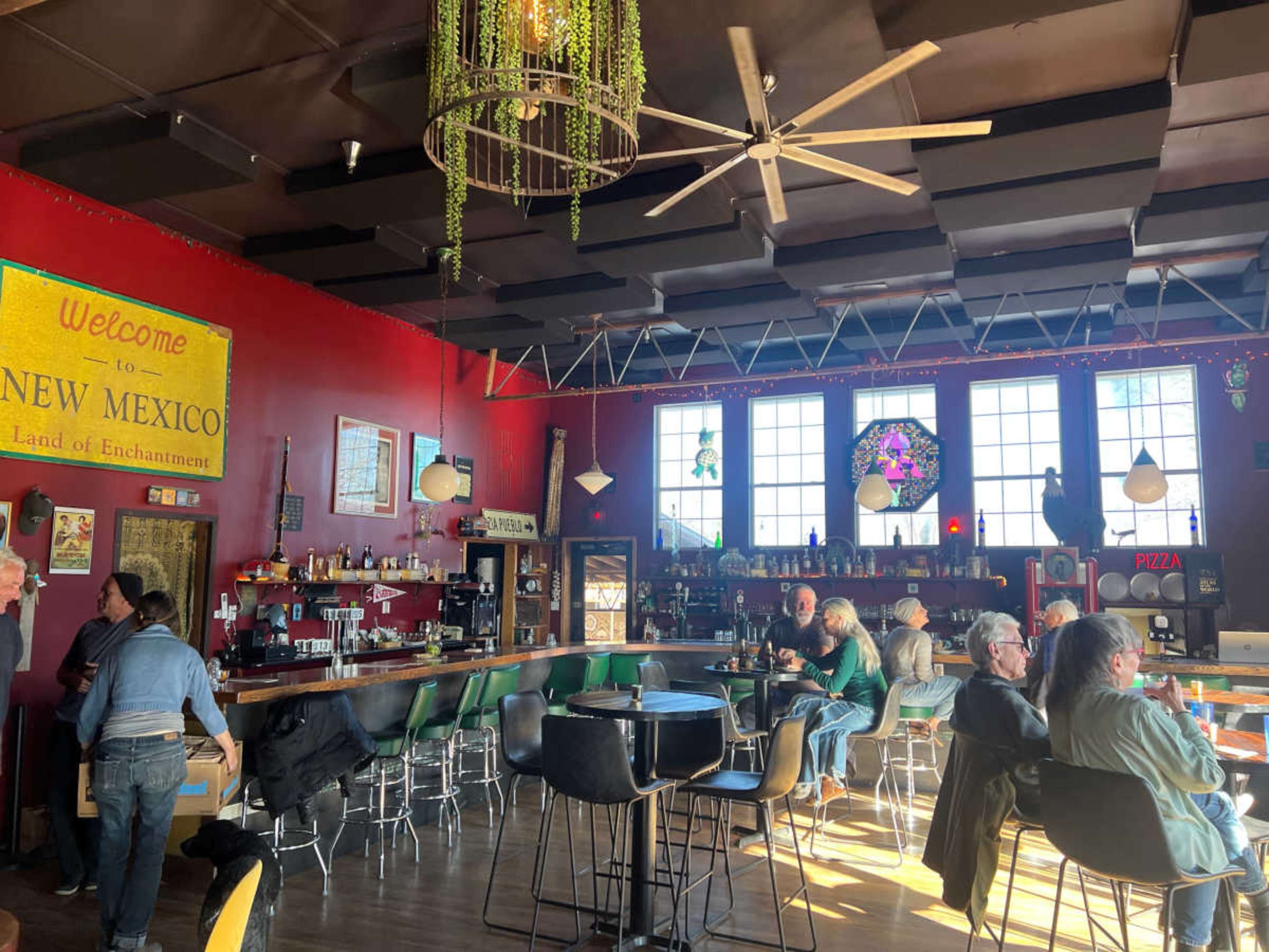 The interior of a bar features colorful decor, a long wooden counter with stools, and patrons seated at tables in a lively atmosphere.