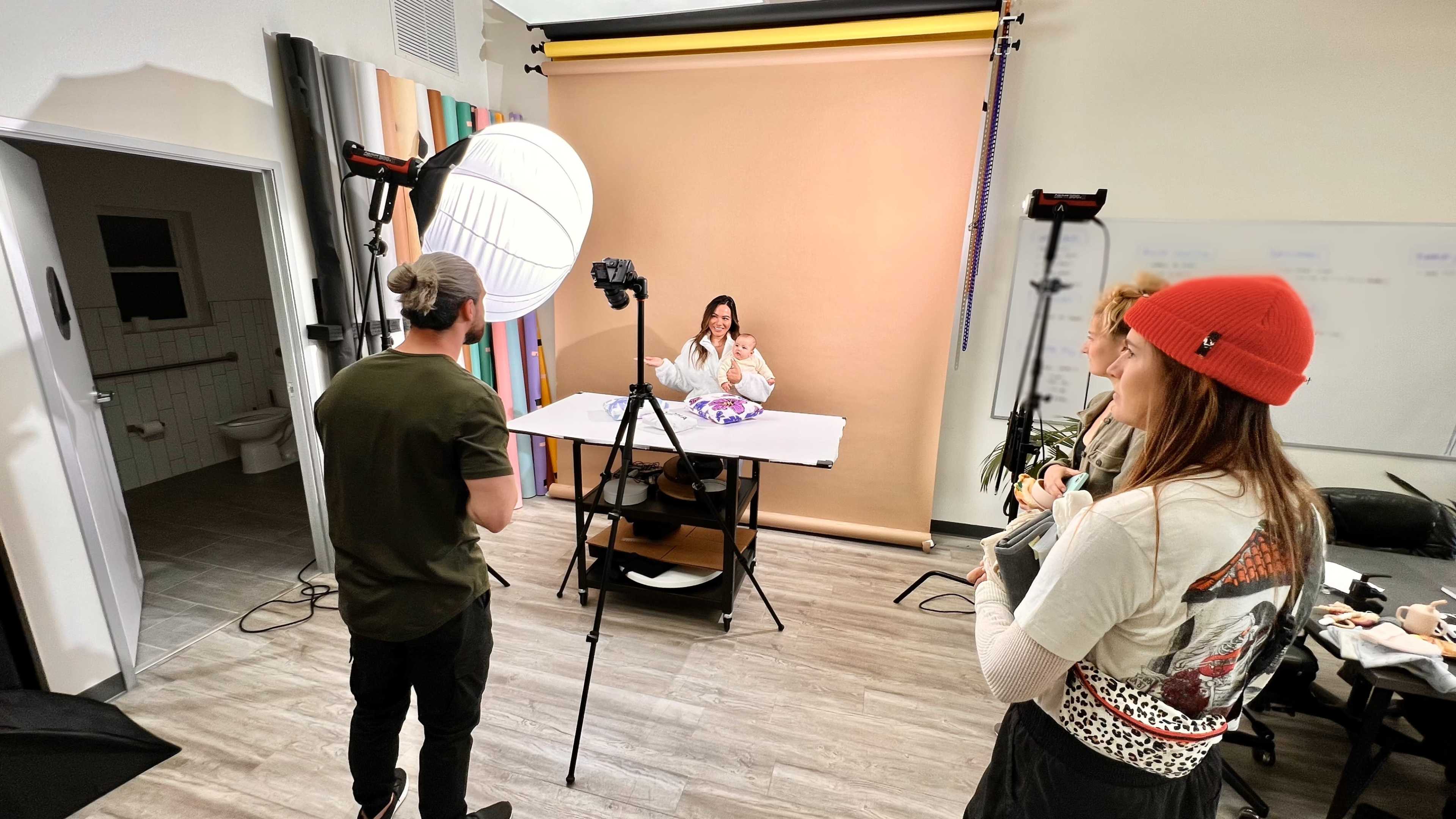 A woman sits at a table in front of a camera, while two people stand nearby, one holding a notebook and another adjusting a light.