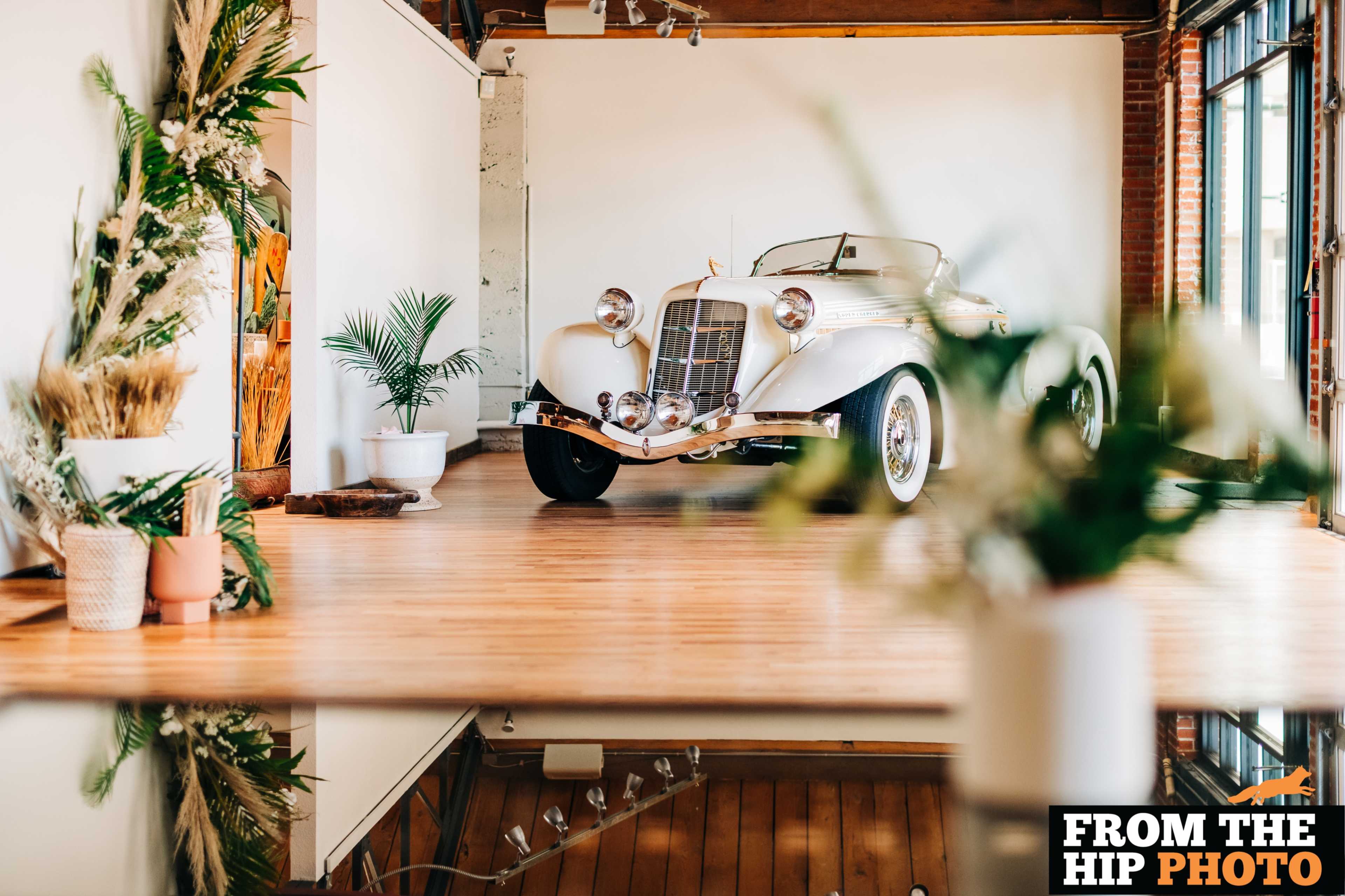 A vintage convertible car is prominently displayed on a polished wooden floor in an artfully designed interior space with plants and decorative elements.