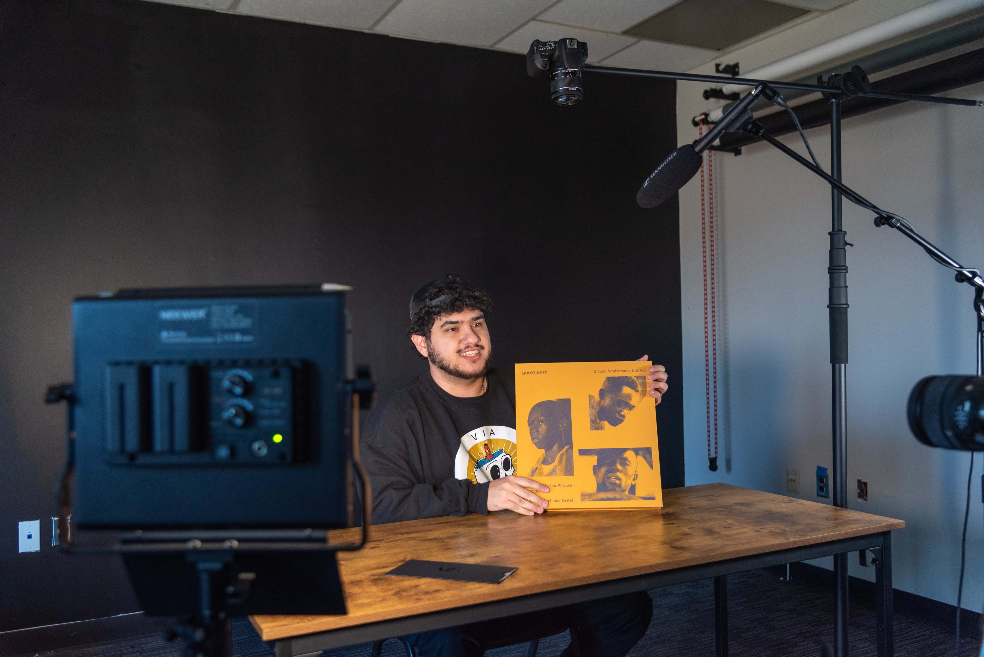 A person holds up a large orange booklet while sitting at a wooden table in a studio setting with video equipment.