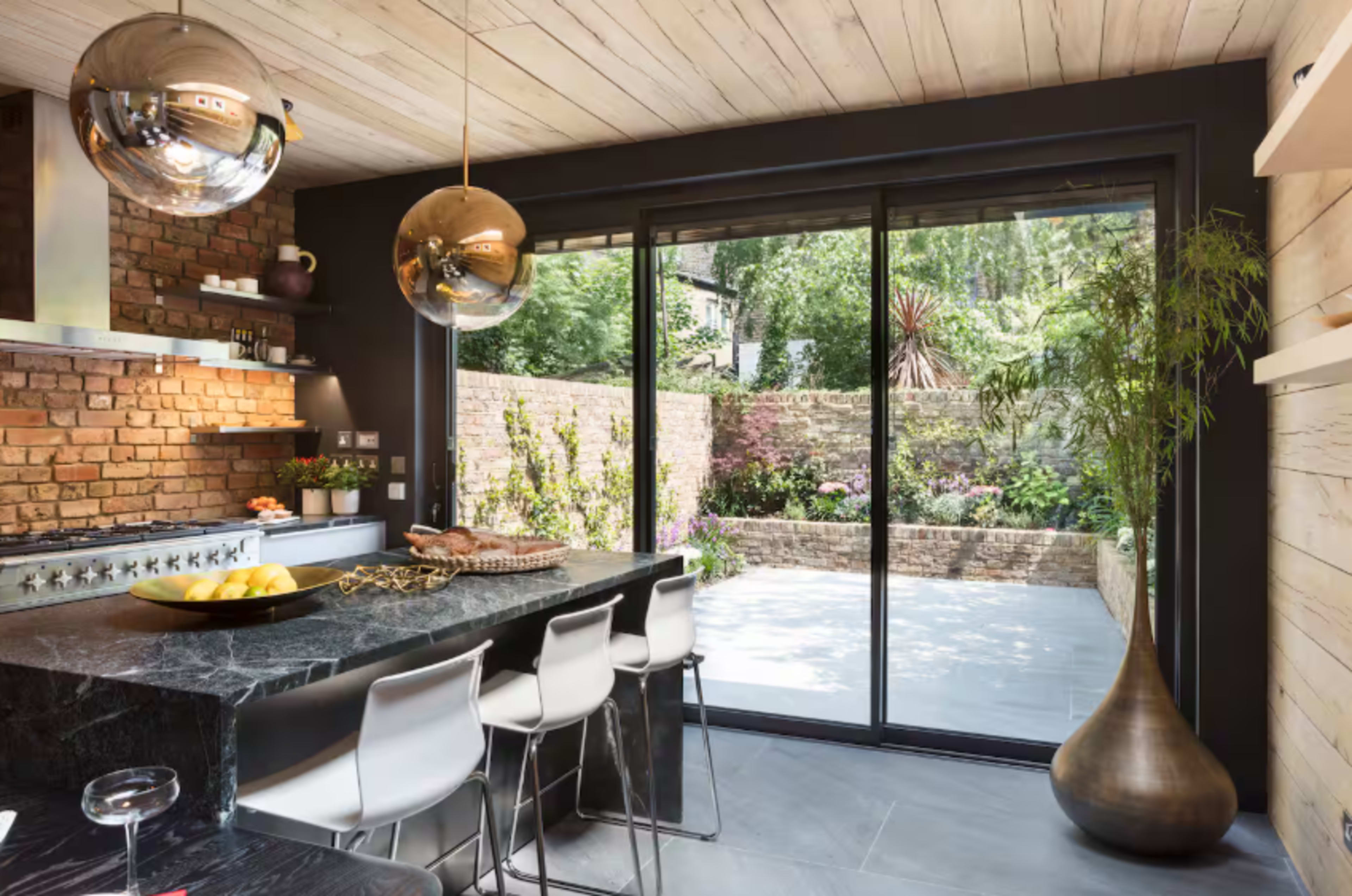 A modern kitchen features a marble island with bar stools, pendant lights, and large windows overlooking a landscaped garden.