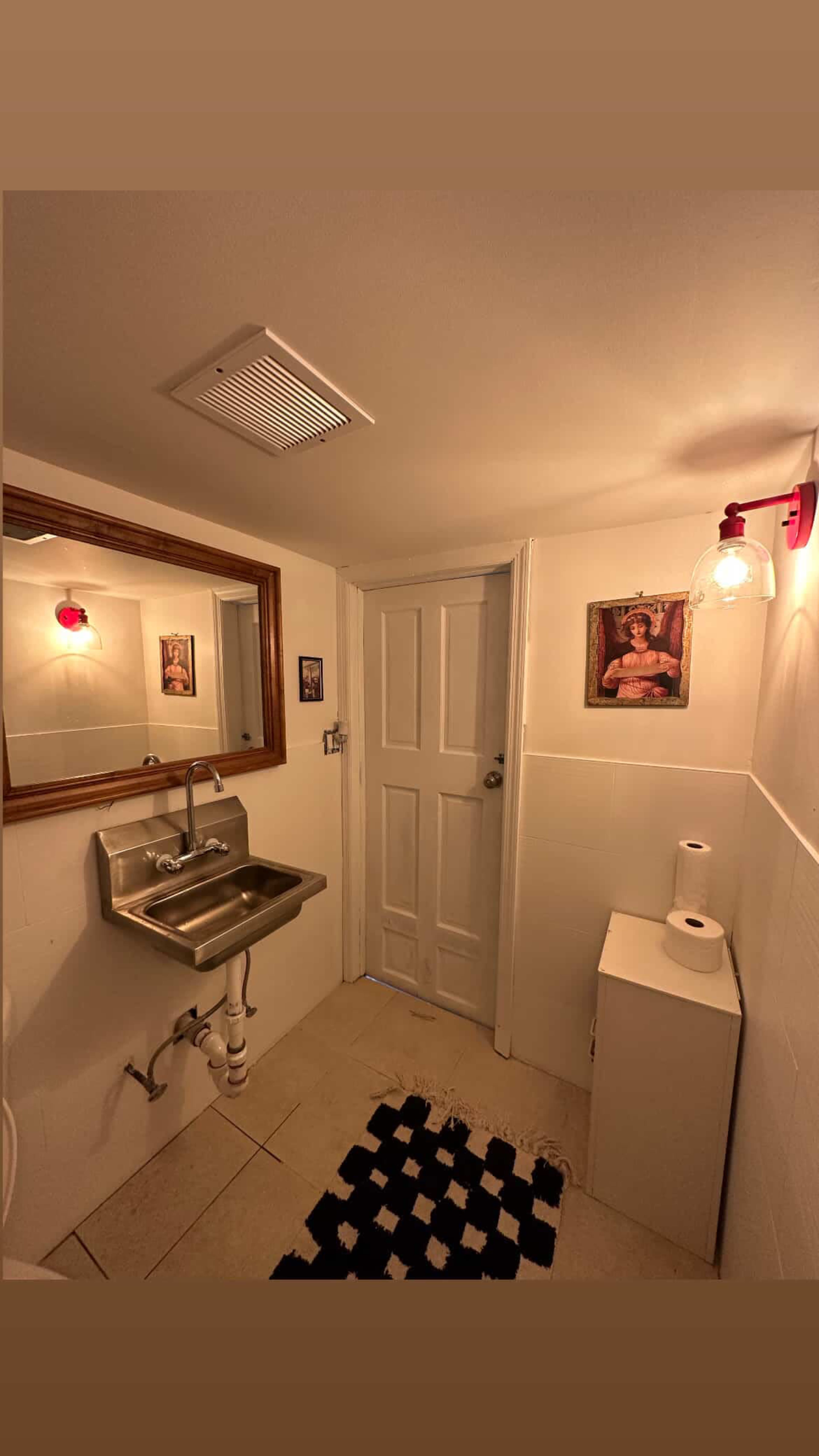 The image shows a small bathroom featuring a sink with a mirror above it, a door, a decorative wall light, and a white toilet beside a checkered rug on the floor.