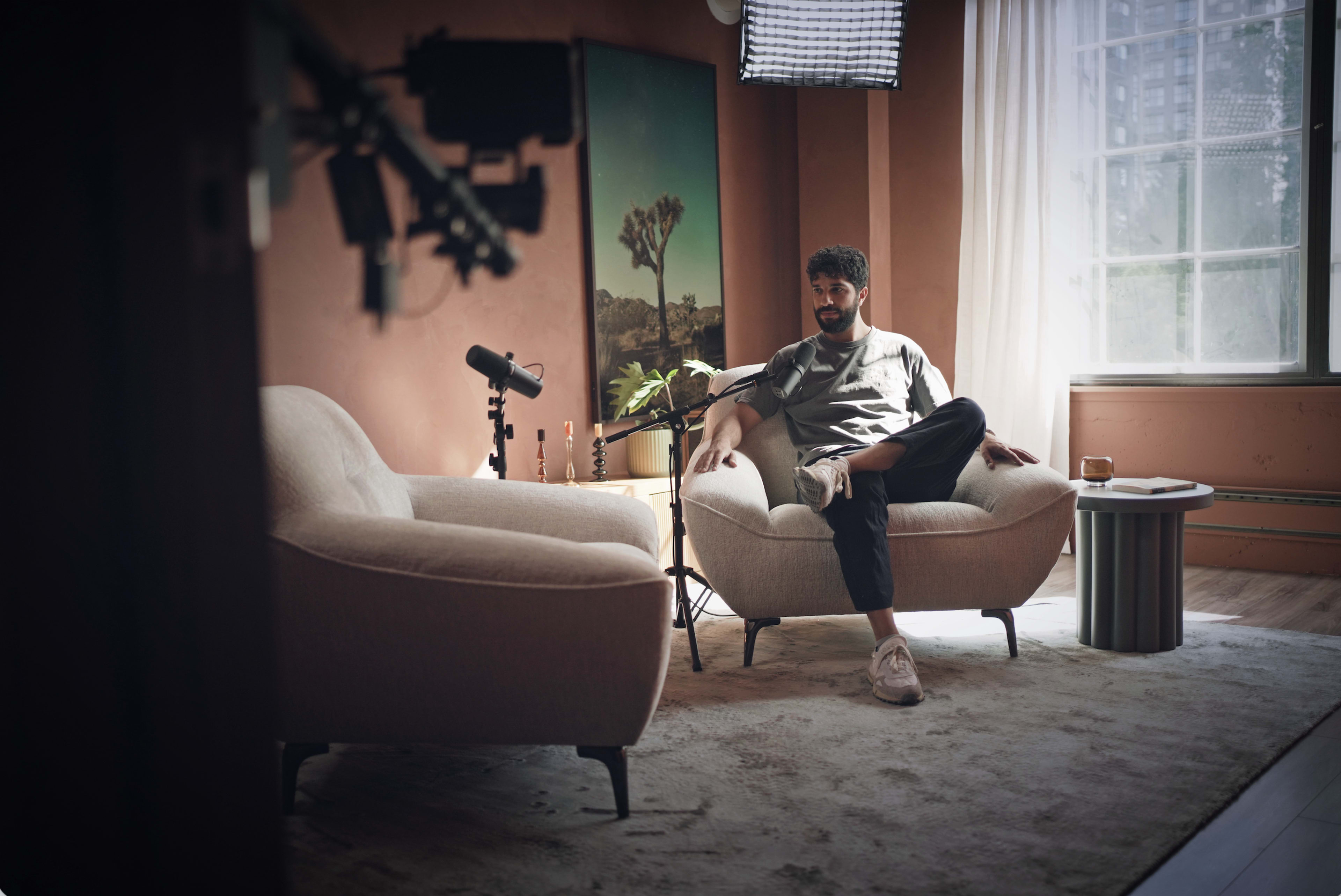 A man sits on a light-colored sofa in a warmly lit room with an art piece on the wall and a small table nearby.