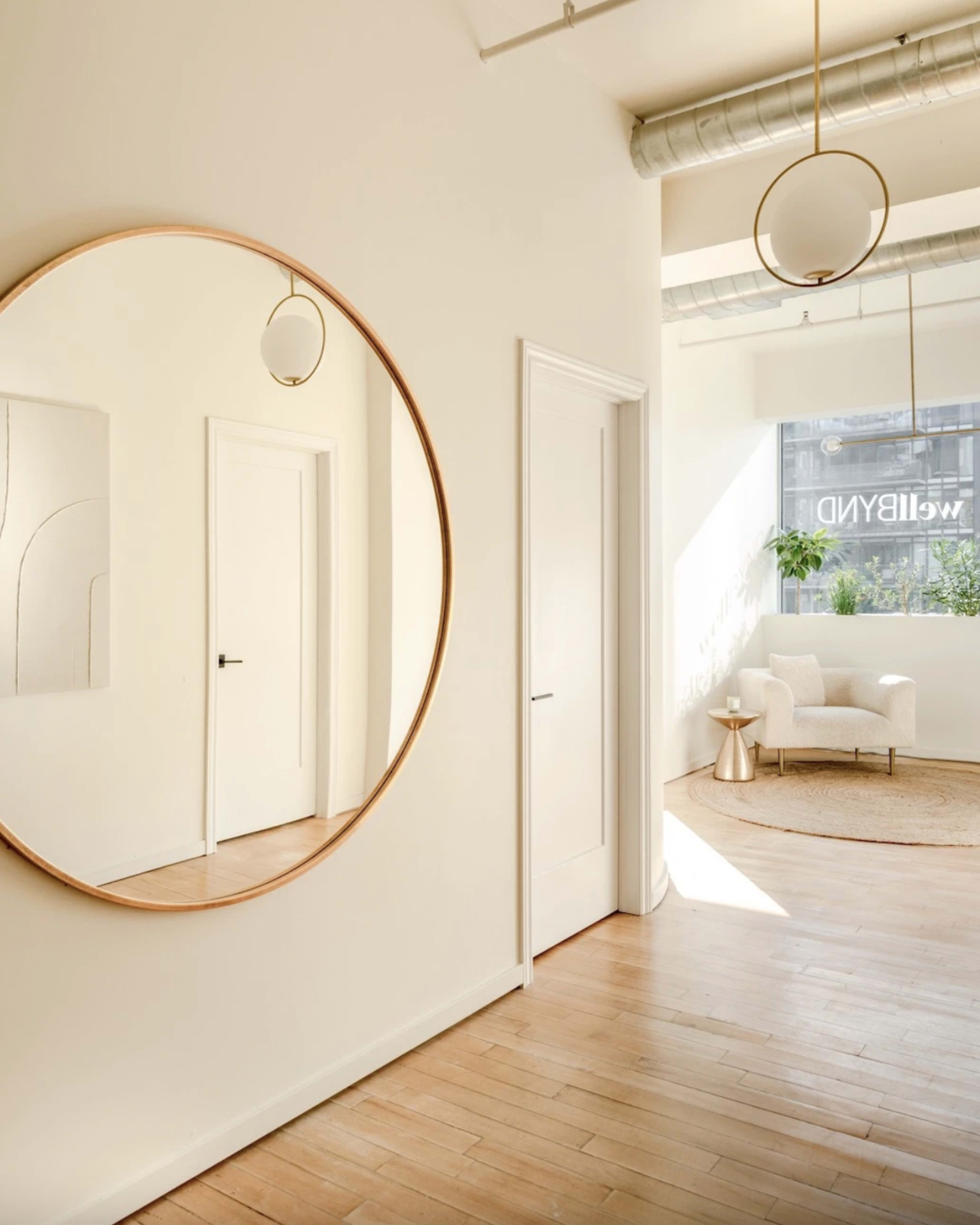 A large circular mirror reflects a bright, minimalist hallway leading to a cozy seating area with a chair and plant.