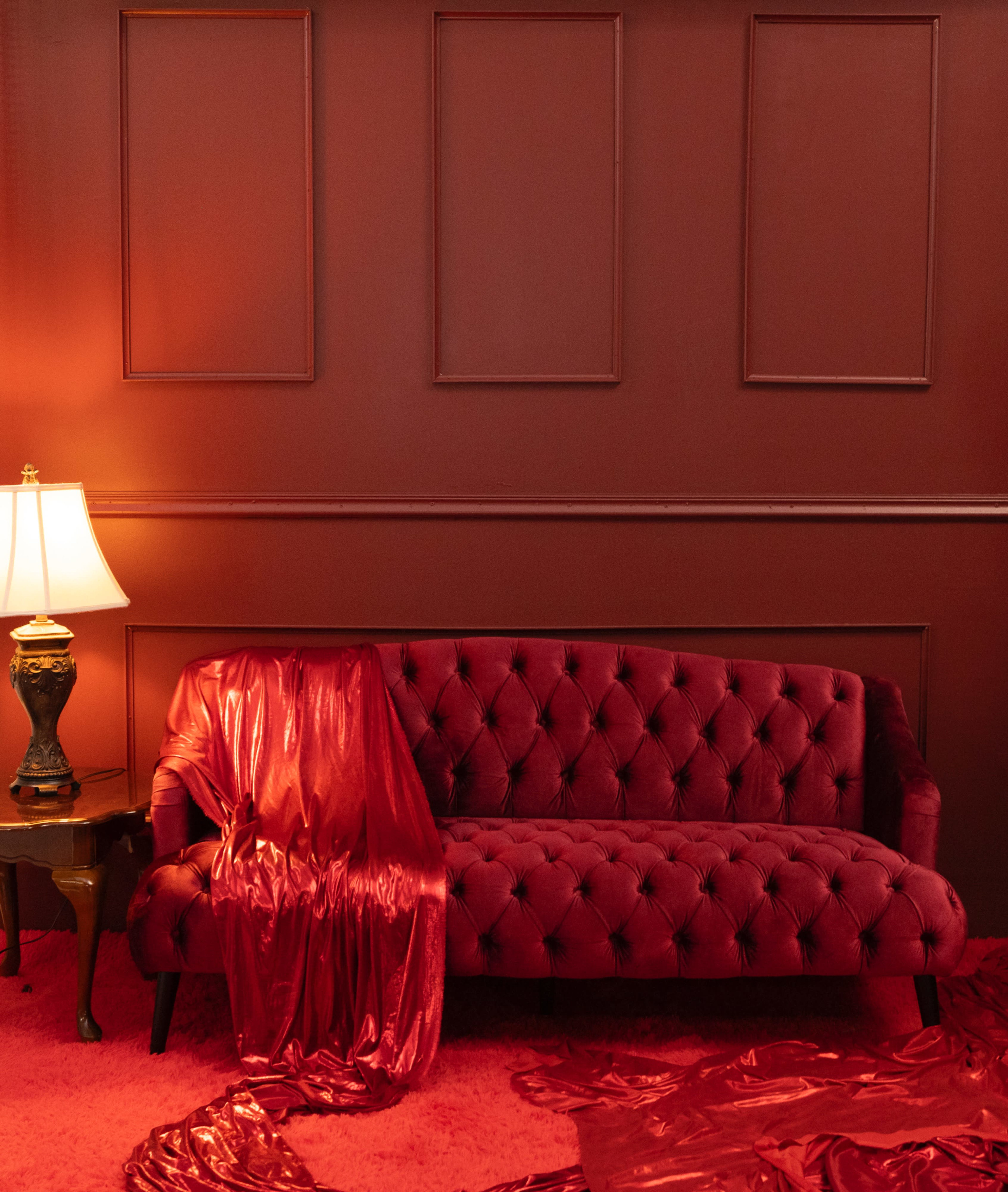A vintage red sofa is positioned against a crimson wall, accompanied by a lamp and draped fabric of similar hues.
