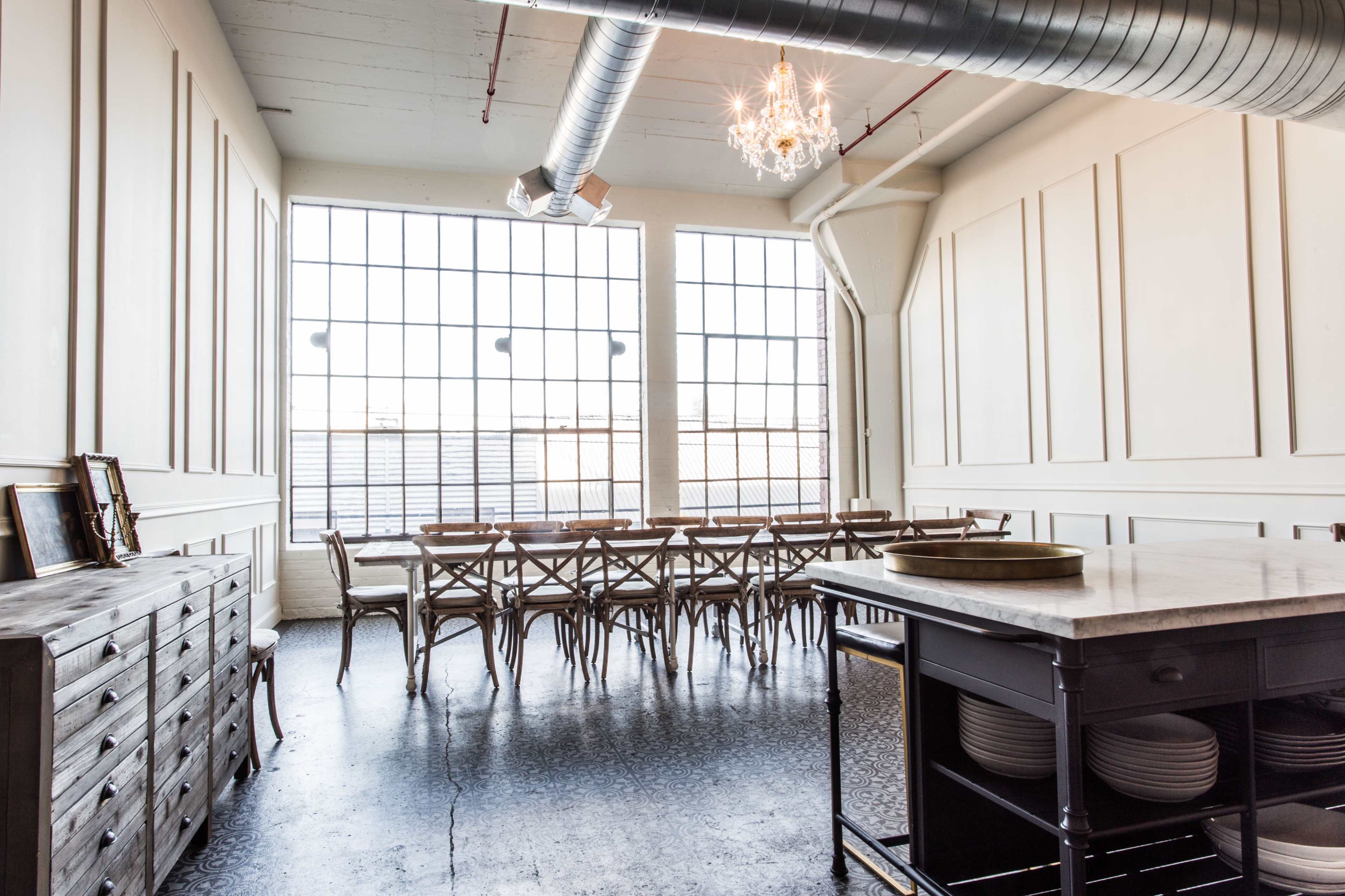 A spacious room with large windows, a long wooden table surrounded by chairs, and a buffet-style counter with plates.