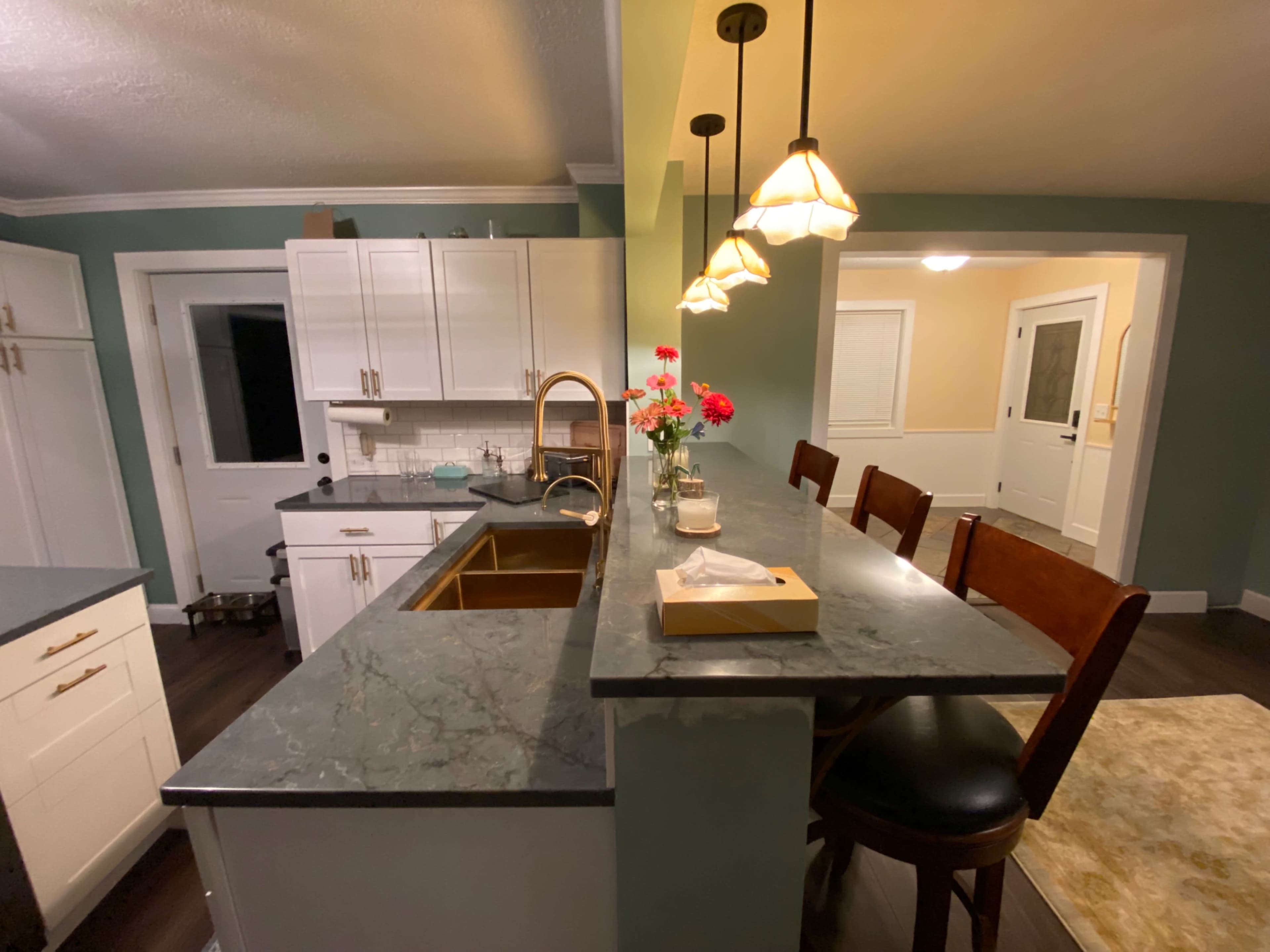 The image shows a modern kitchen featuring a marble countertop, wooden bar stools, and a vase of flowers on the island.