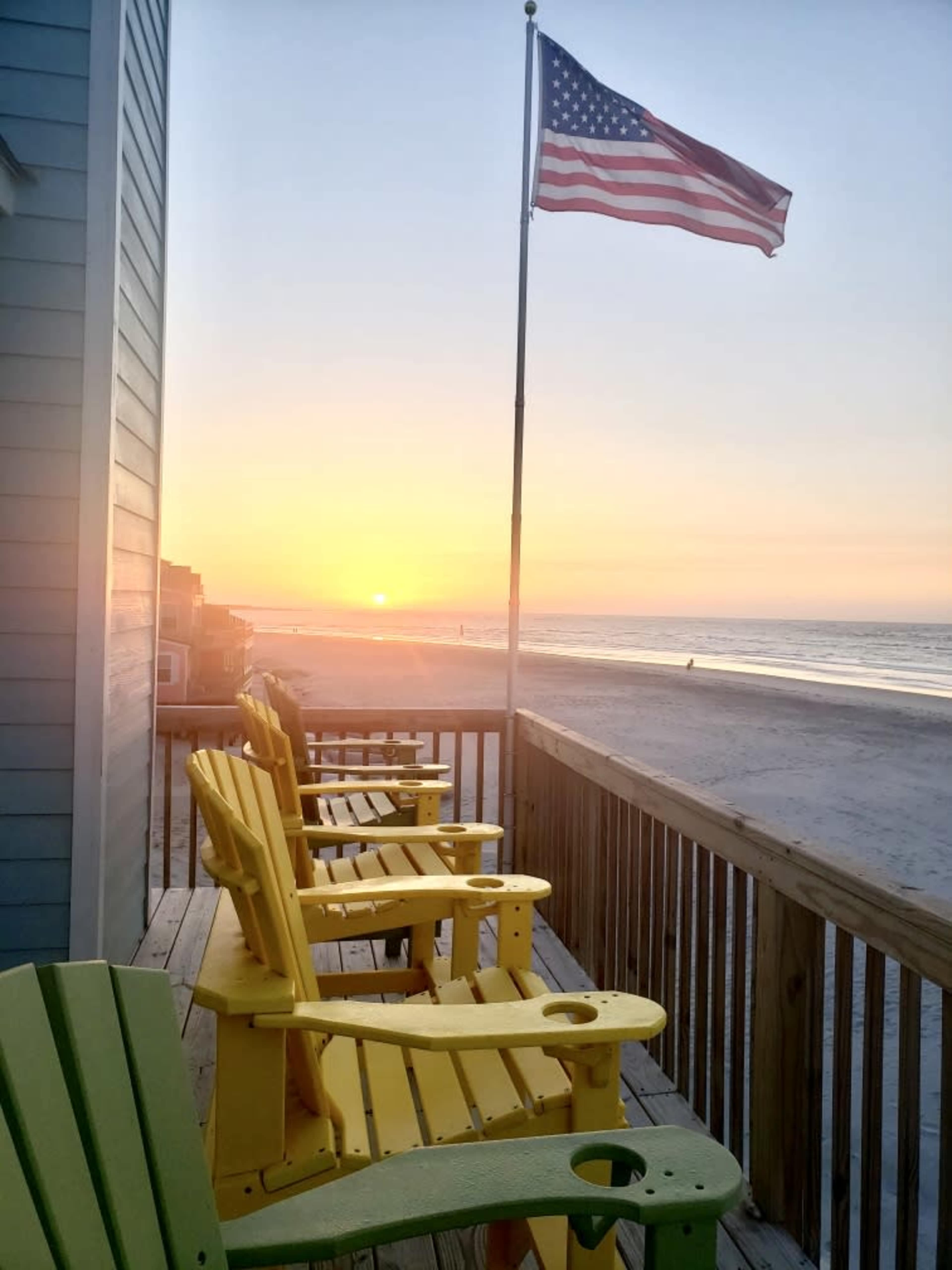 A row of yellow adirondack chairs lines a wooden deck overlooking a beach at sunrise, with an American flag fluttering in the breeze.