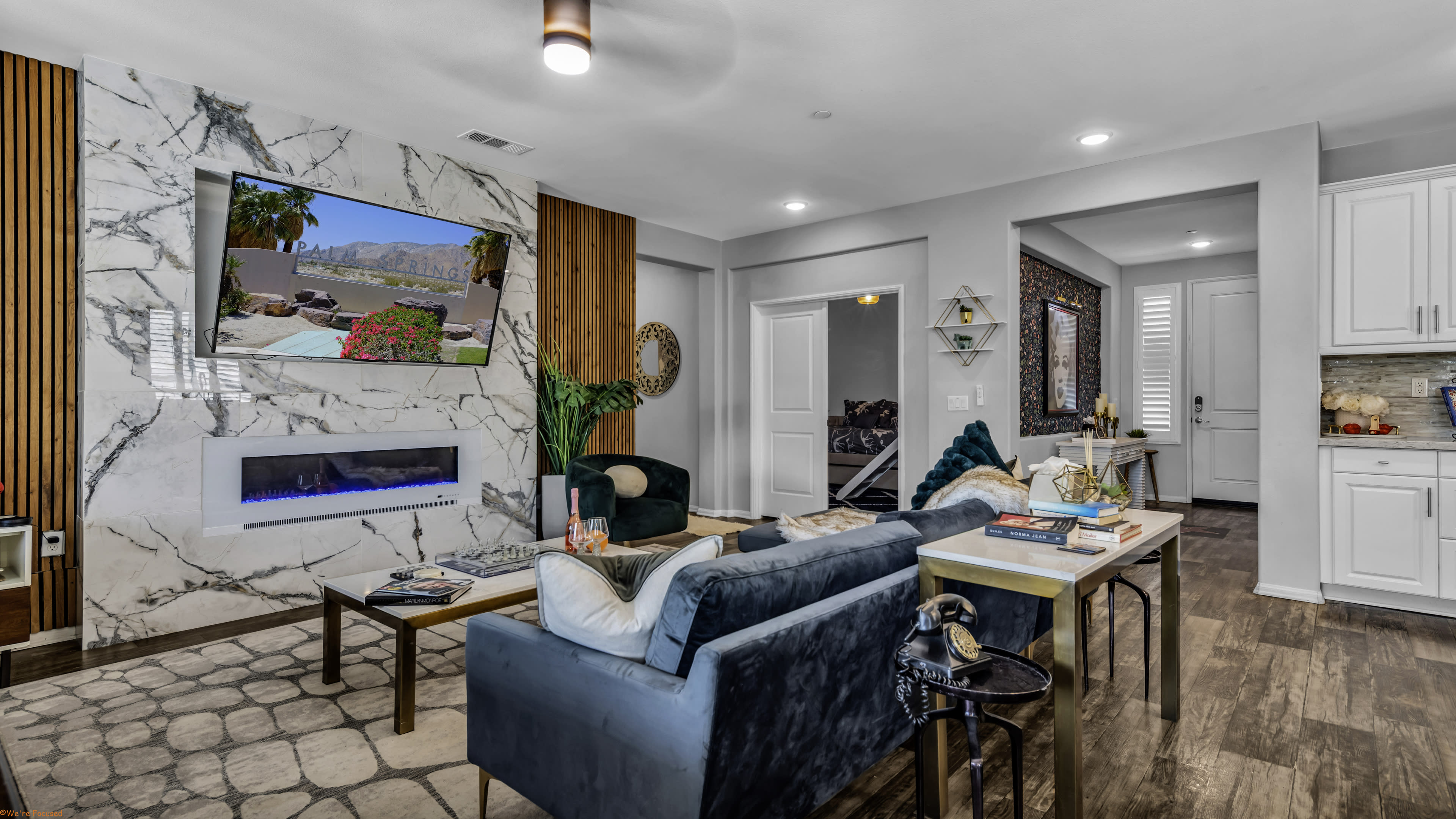 The living room features modern furniture, a large wall-mounted television, and a stone fireplace with a minimalist style, leading to an adjoining kitchen area.