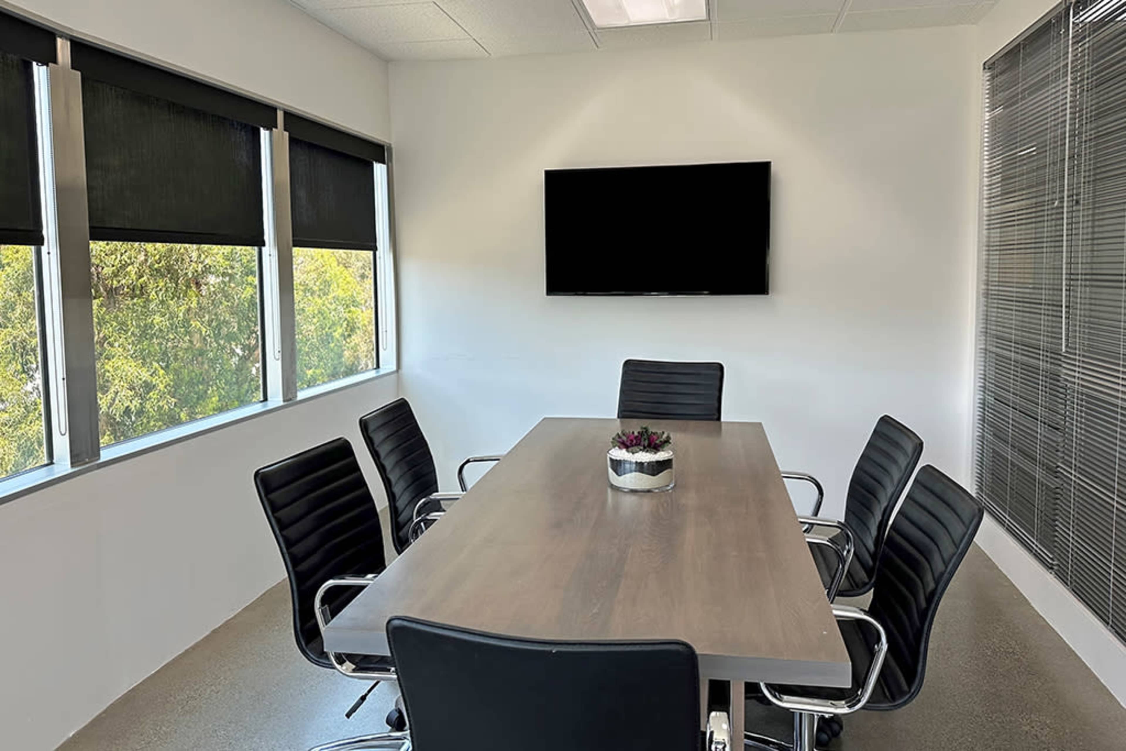 A modern conference room features a long wooden table surrounded by six black chairs, with large windows and a wall-mounted television.