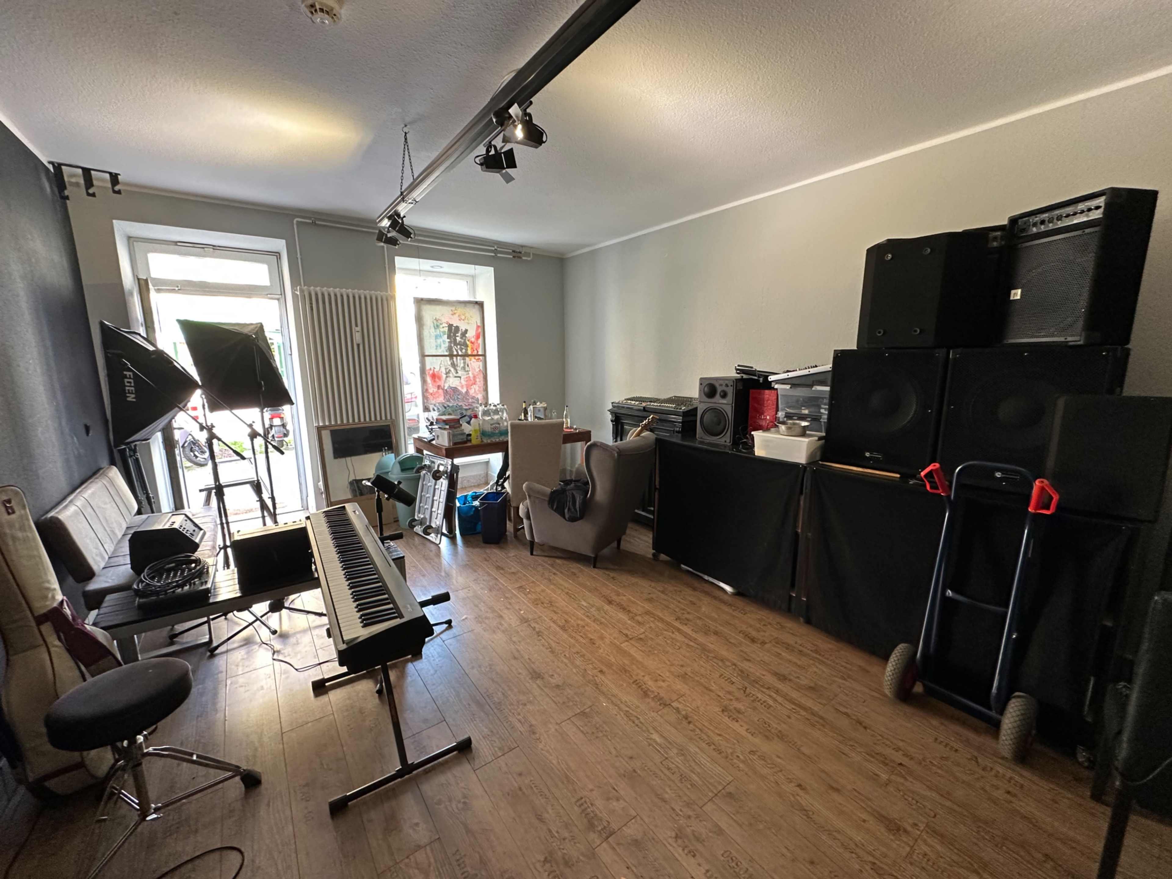 The image shows a music rehearsal space with a keyboard, various speakers stacked in the corner, a chair, and a distinct wooden floor.