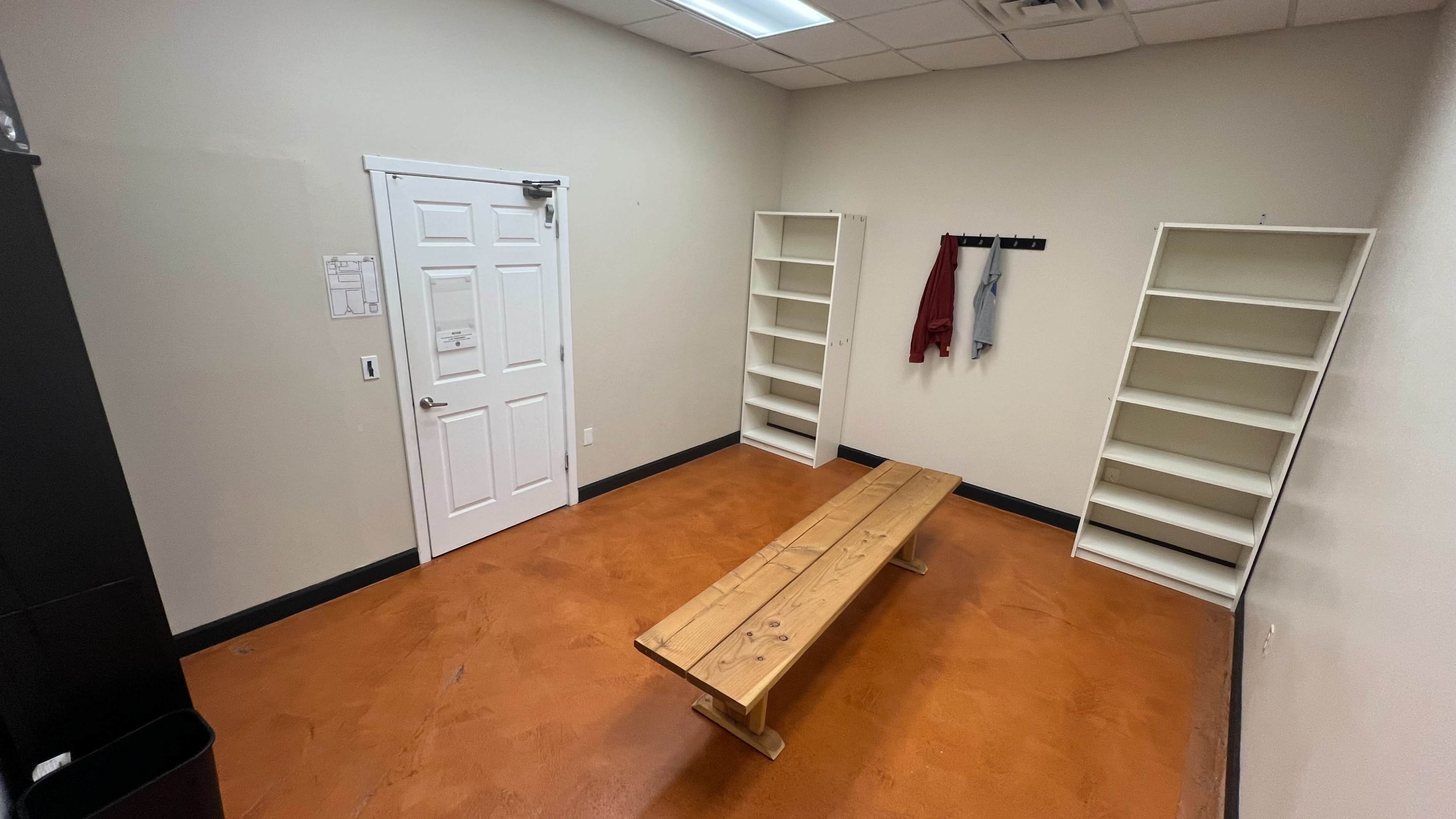 The image shows a small, empty room with an orange floor, a wooden bench in the center, a door on one wall, and two empty shelves on either side.