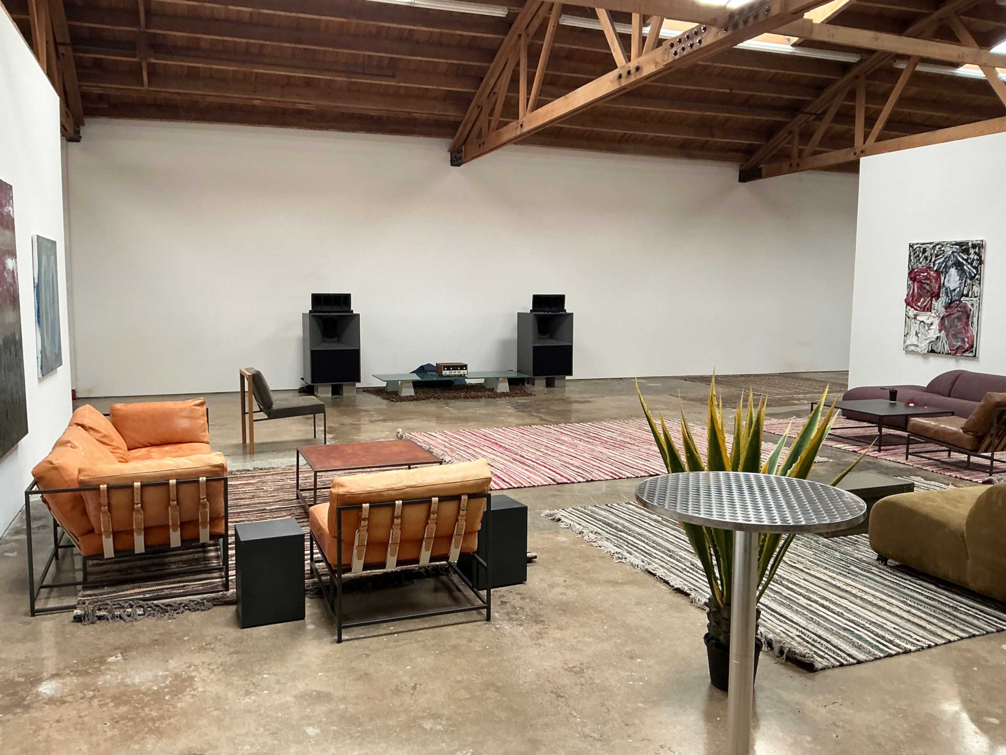 The image shows a spacious art gallery with minimalistic furniture, including orange and brown seating, a coffee table, and decorative rugs on a polished concrete floor.