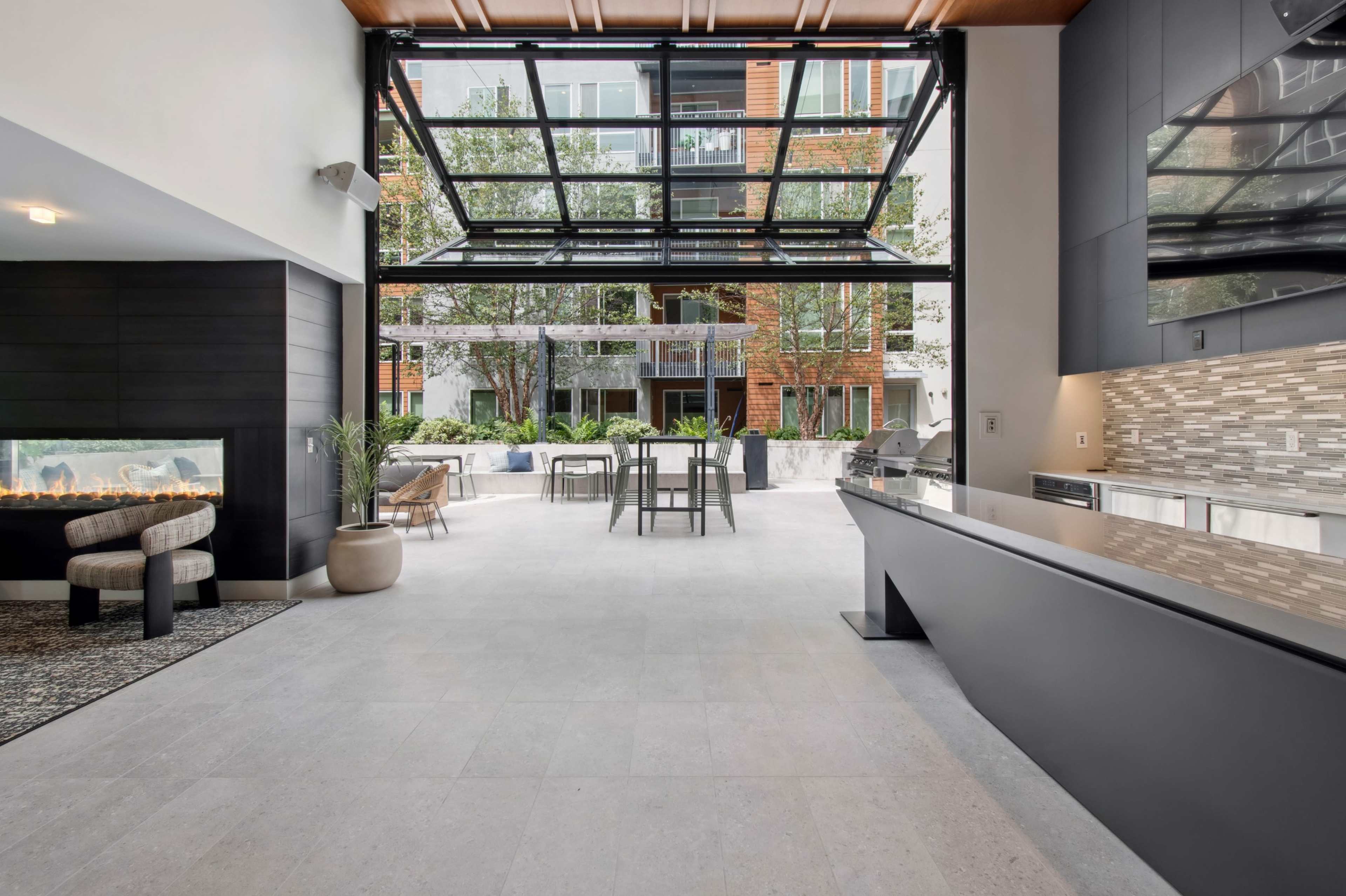 The image shows a modern indoor space that opens to an outdoor area, featuring a kitchen with sleek countertops, a fireplace, and a dining table surrounded by greenery.