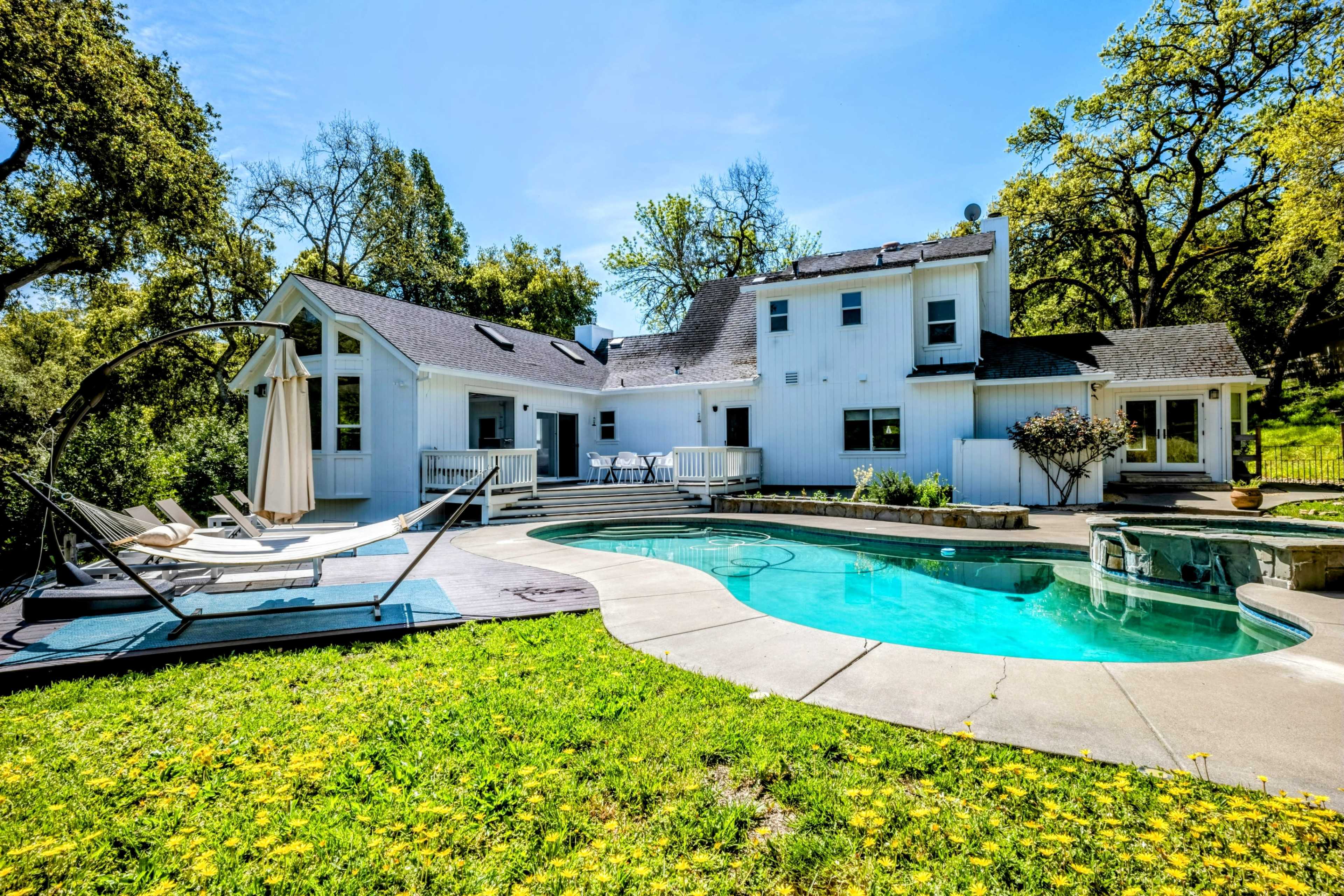 A white house with a swimming pool and lounge chairs in a grassy backyard surrounded by trees.