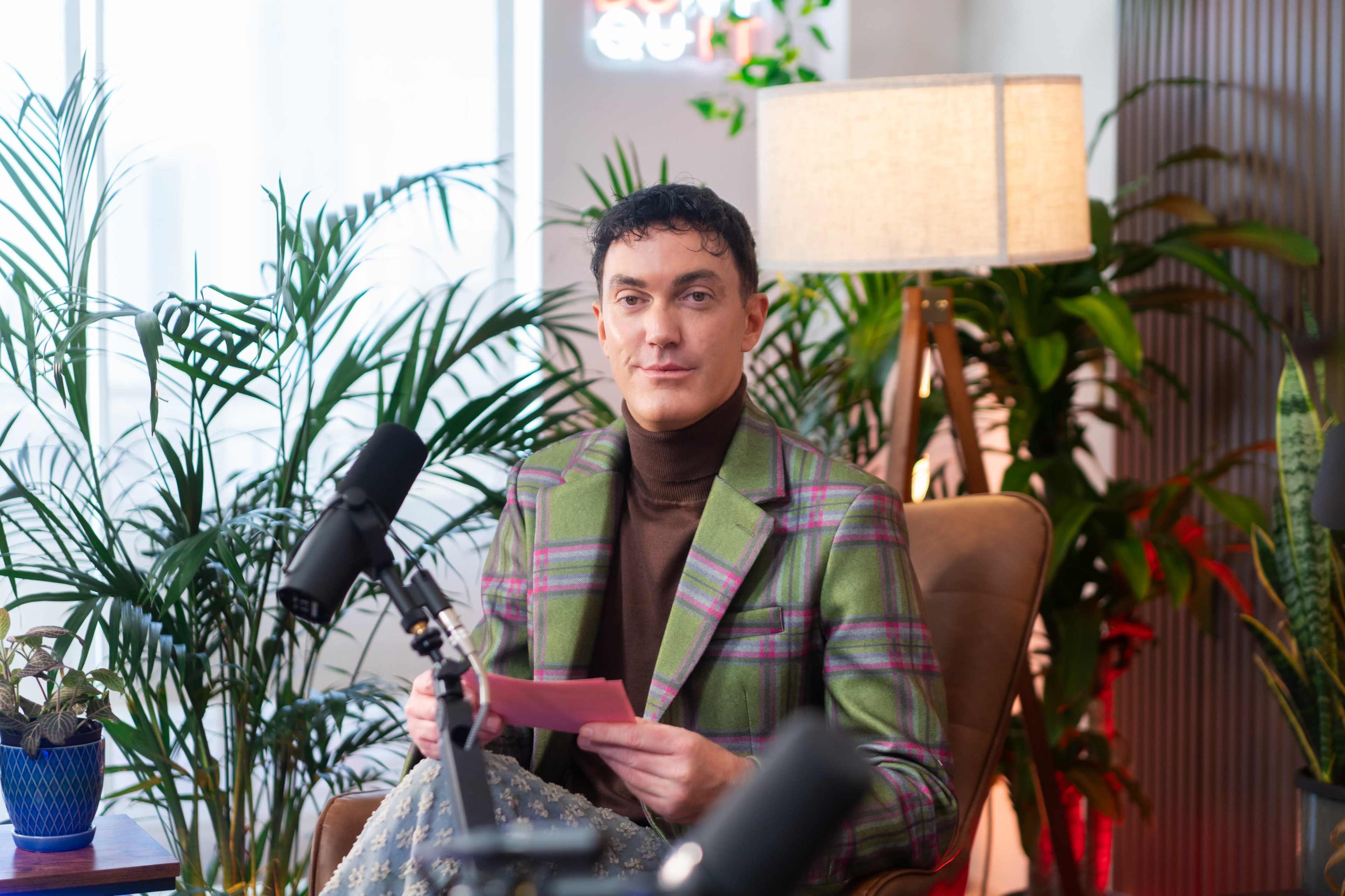 A person sits in a chair holding a paper, surrounded by plants and a lamp in a well-lit room.