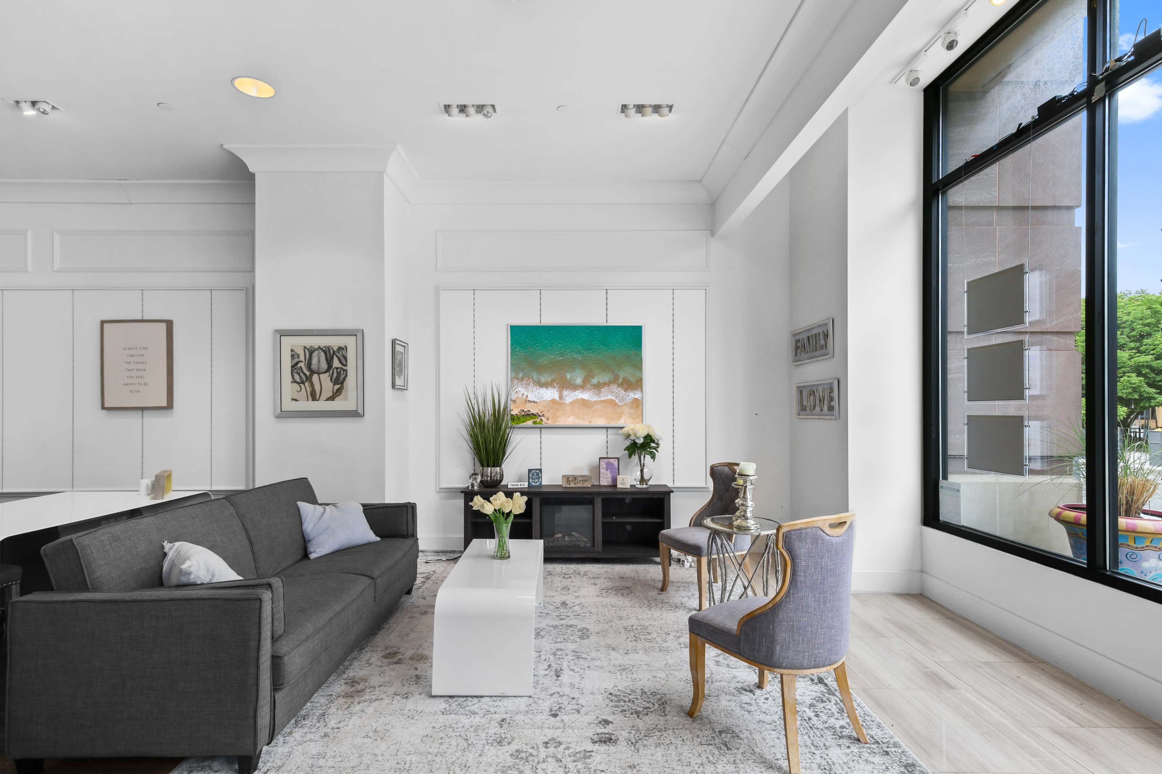 A modern living room features a gray sofa, white coffee table, and large windows with a view, highlighted by an abstract painting and plants.