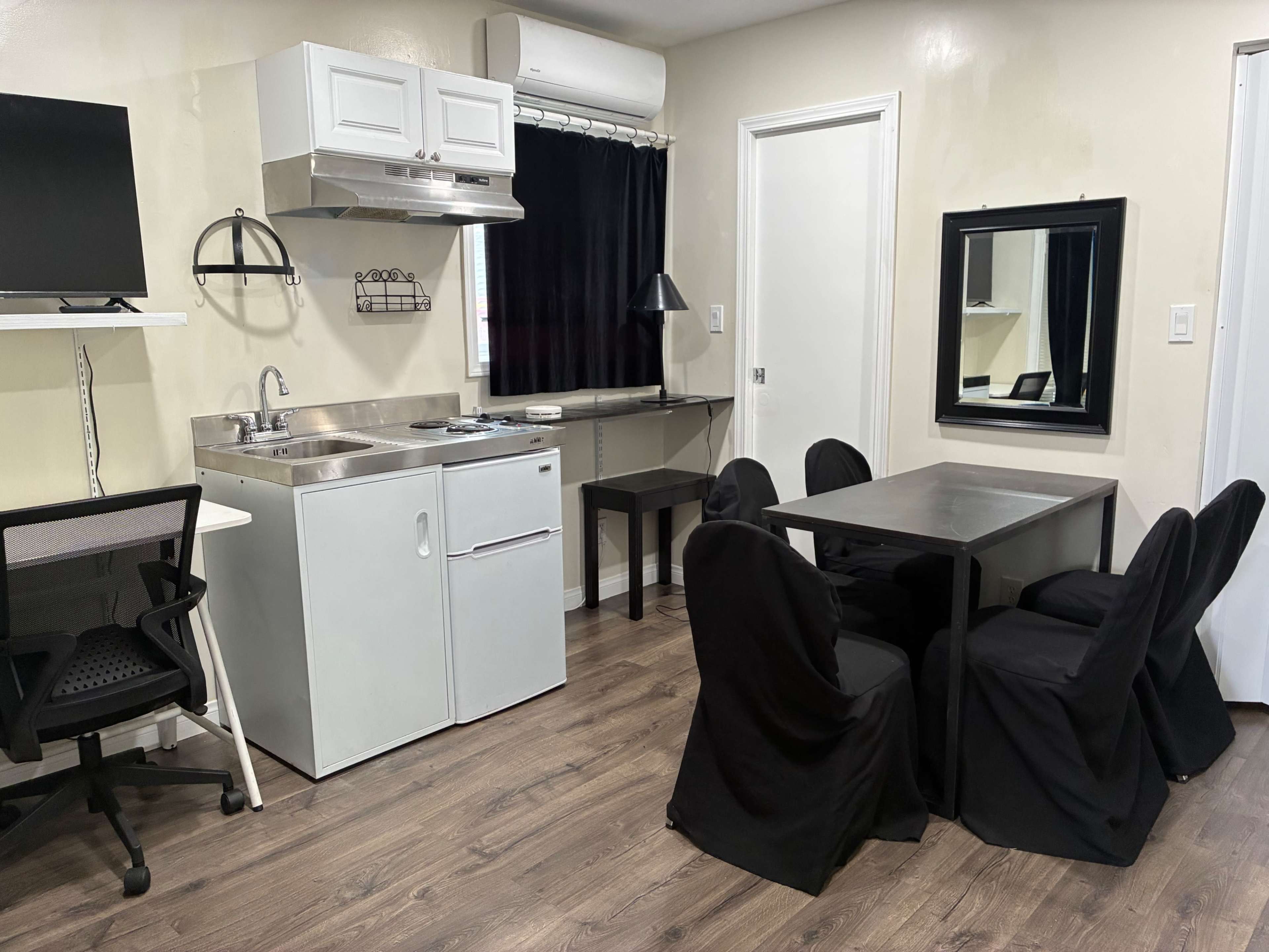 The image shows a compact kitchen and dining area featuring a small table with six chairs, a sink, a stovetop, and a wall-mounted television.